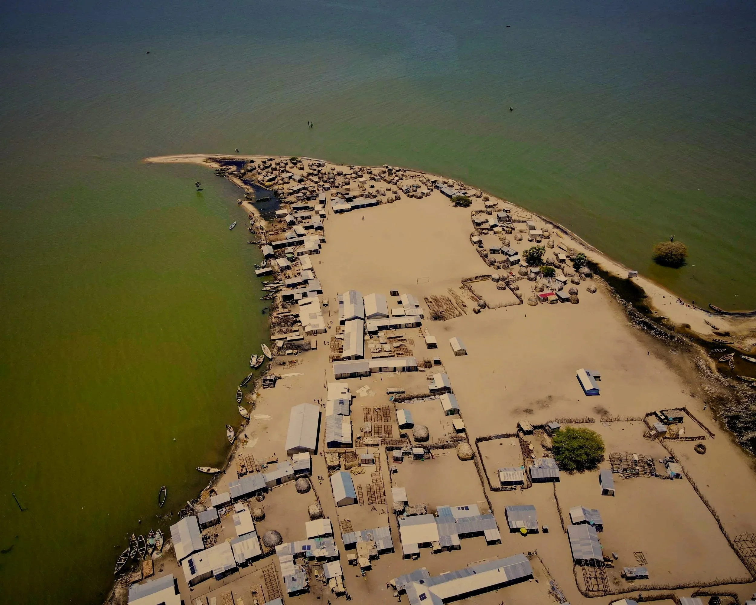 Aerial view of a small coastal village with white buildings and thatched roofs, situated on a narrow strip of sandy land extending into calm green water, with boats docked along the shoreline and a few structures and trees.