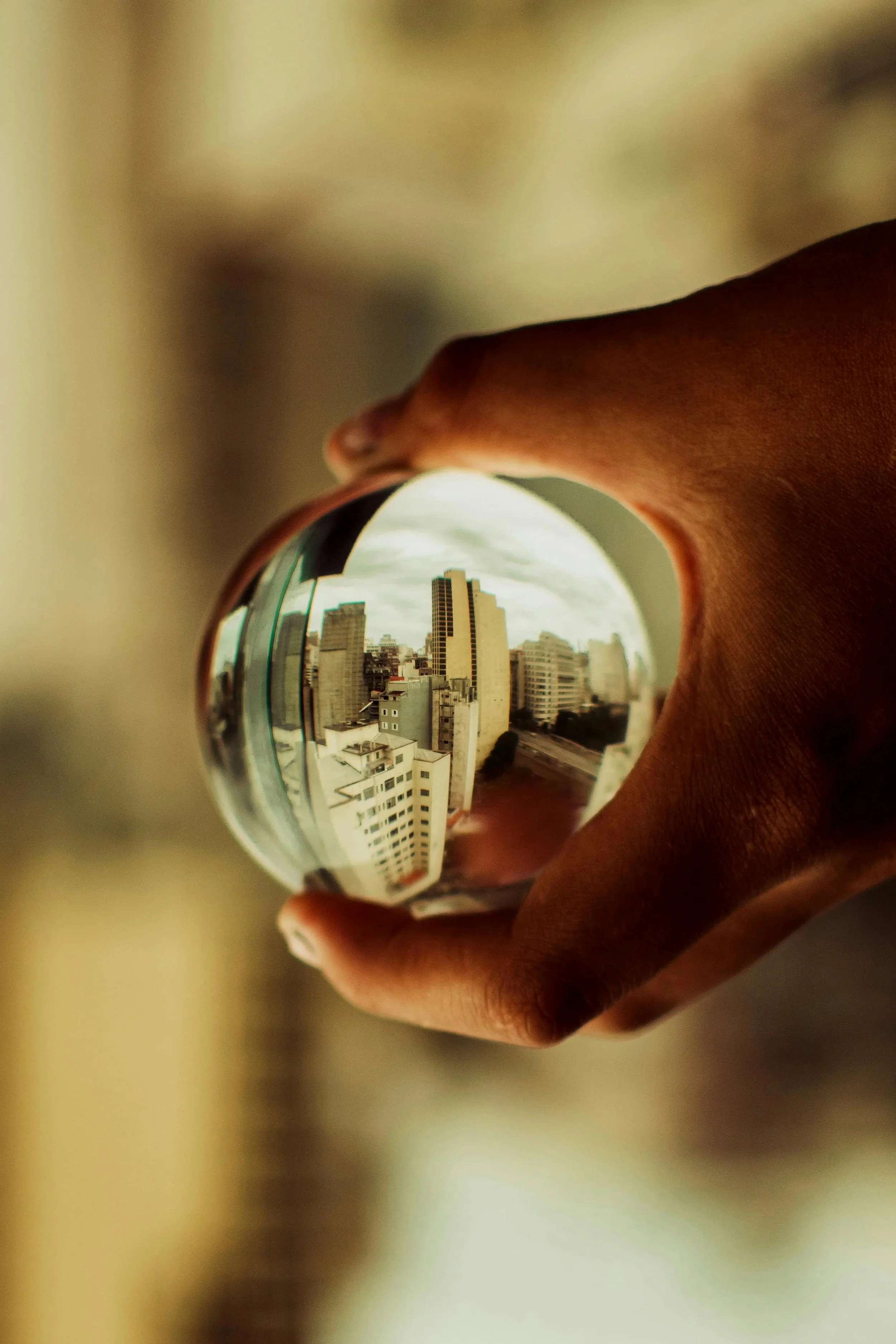 A hand holding a glass sphere that reflects a cityscape of tall buildings and cloudy sky.