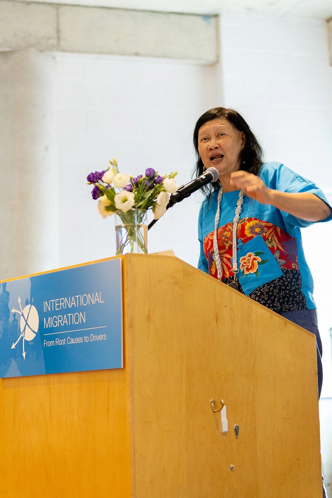 A woman giving a speech at a podium with a sign that reads 'International Migration: From Root Causes to Drivers'. There is a floral arrangement on the podium.