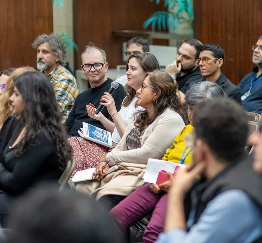 A diverse group of people sitting in a conference or seminar, one woman is holding a microphone and a booklet, and others are listening attentively.