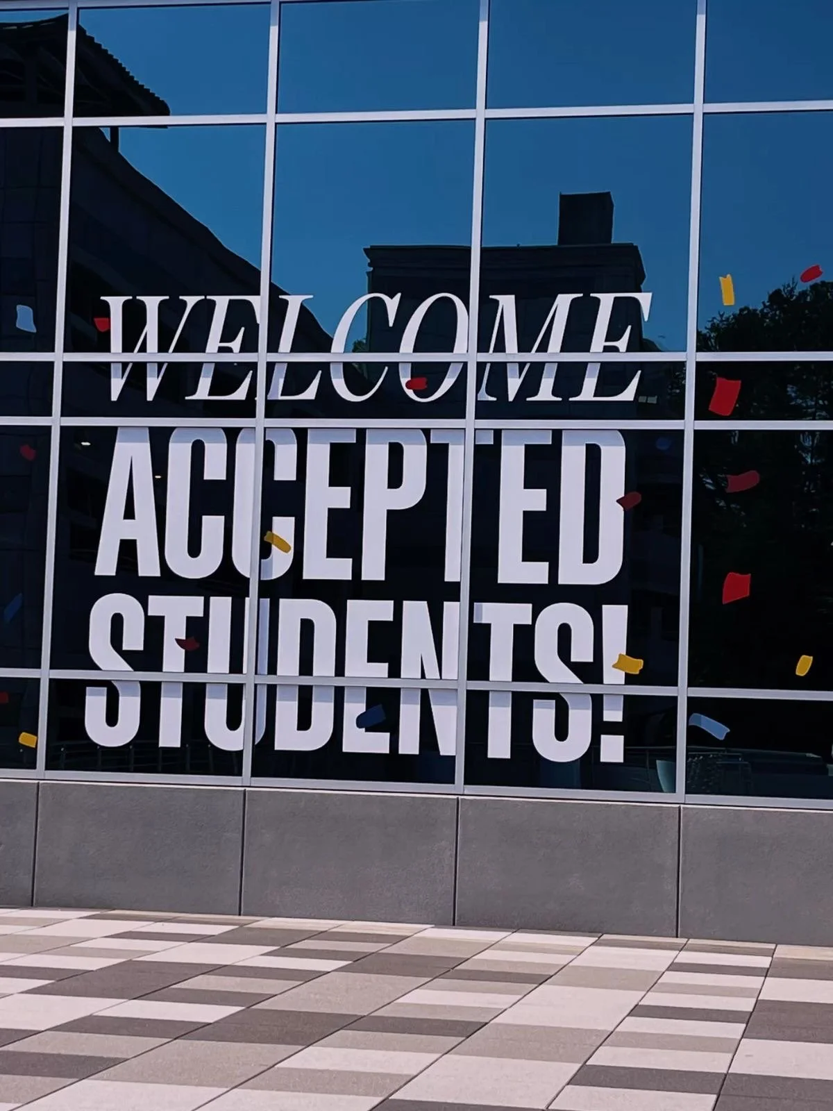 A large glass window with the message 'Welcome Accepted Students!' written on it, surrounded by colorful confetti and reflecting nearby buildings.