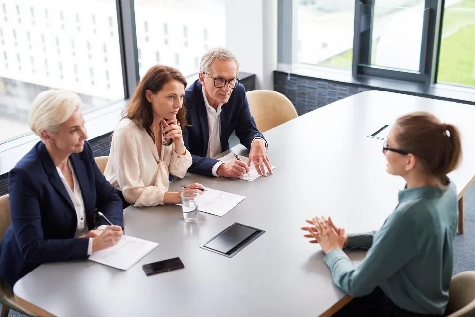 Four people sitting at a conference table in a bright office, engaged in a conversation with a woman on the right side.