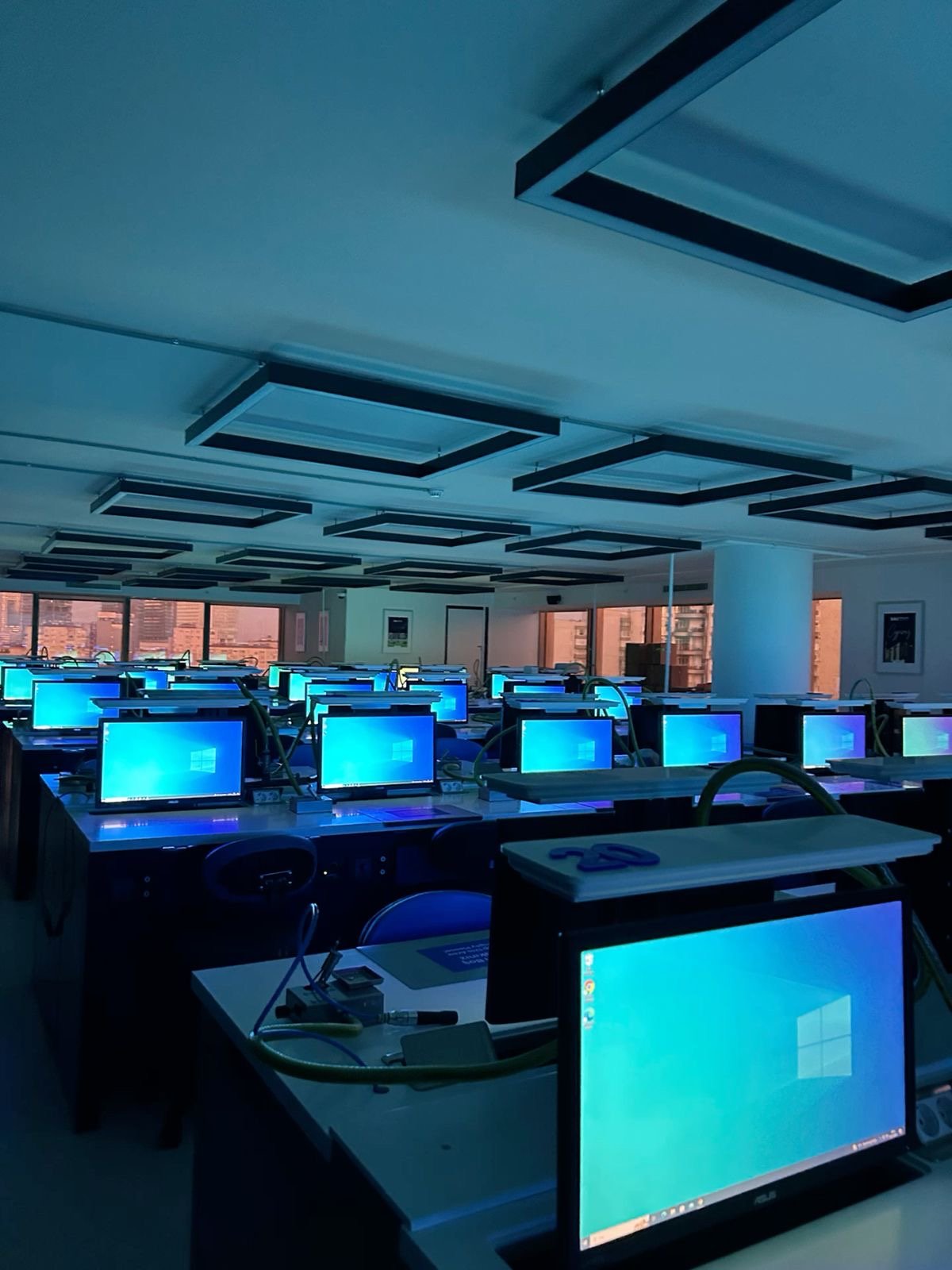 A classroom filled with rows of computers with blue screens, windows showing a city skyline, and ceiling lights.