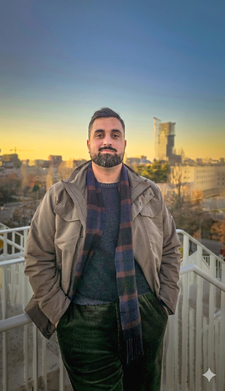 A man with a beard and short dark hair standing outdoors on a balcony during sunset, wearing a brown jacket, a dark sweater, and a blue and red checkered scarf, with a cityscape in the background.