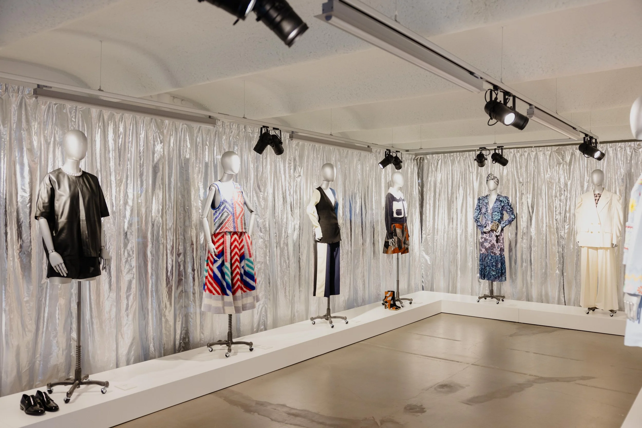 Jane mannequins in colorful and black outfits on display in a fashion boutique, with a metallic silver curtain backdrop and track lighting