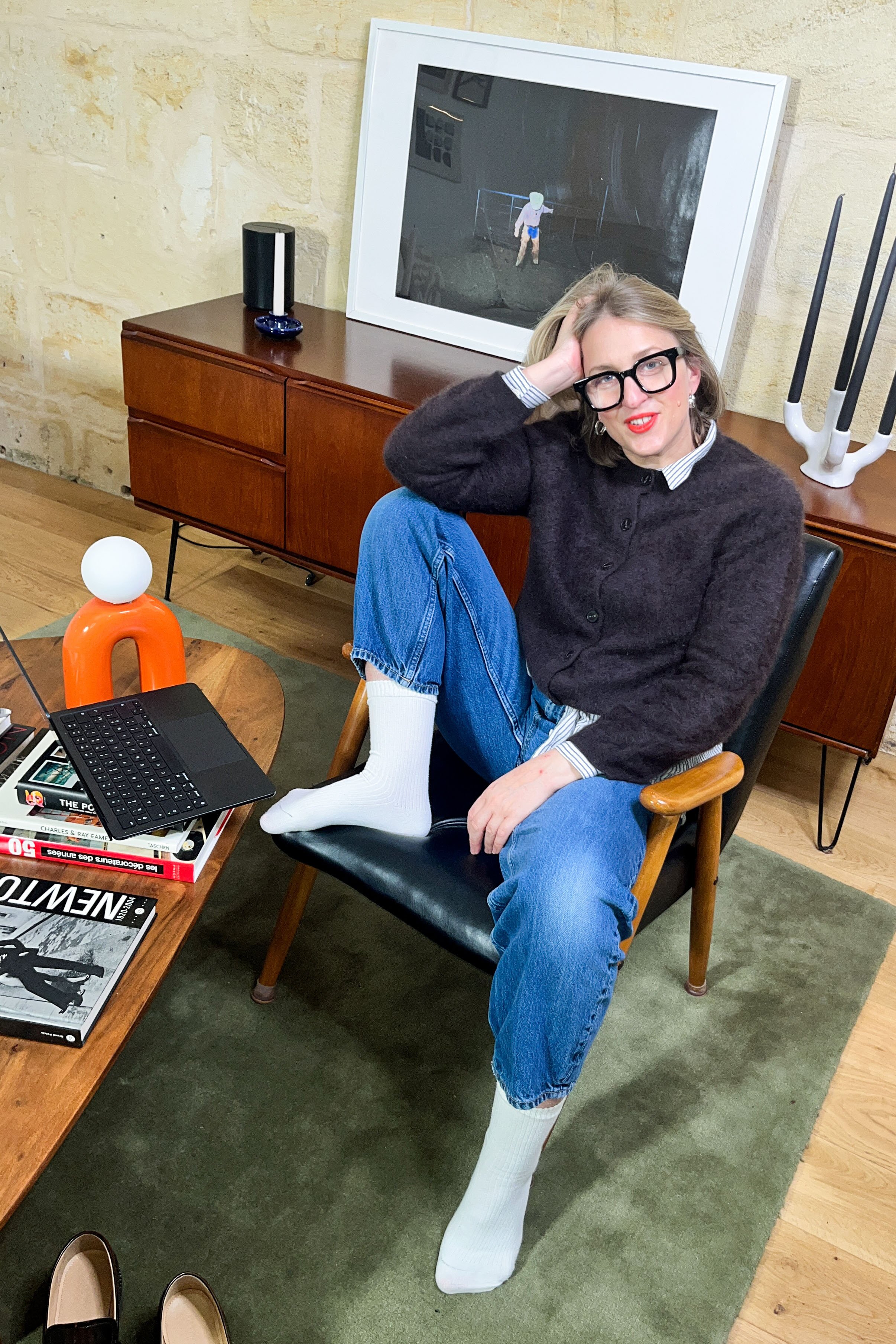 A woman sitting in a black chair with her legs crossed, wearing white socks, jeans, a striped shirt, and a dark cardigan, with glasses and red lipstick, in a room with a wooden floor and a green rug, a mid-century modern wooden sideboard, a framed picture of a tennis player, and a side table with books, decorative objects, and a computer.