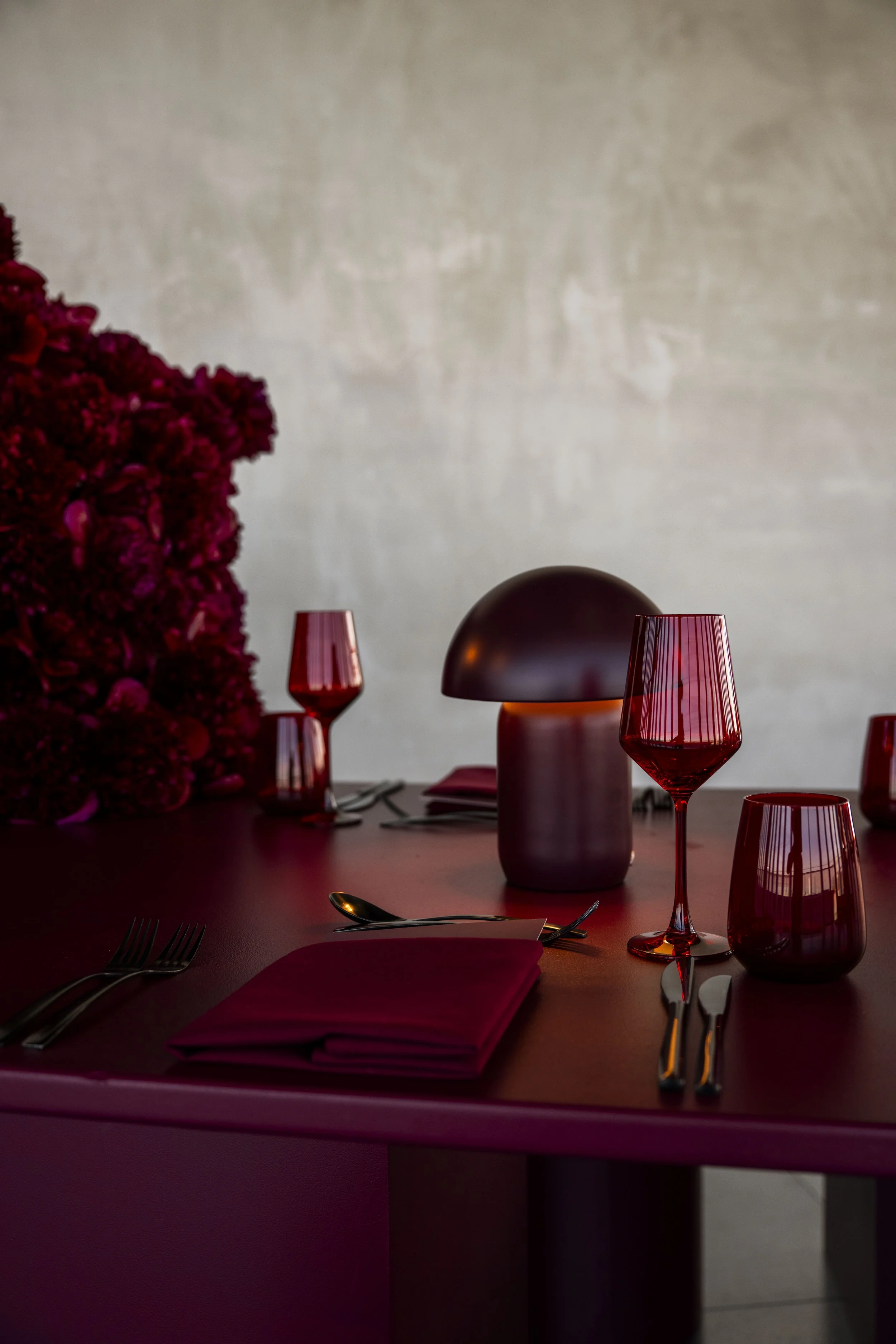 Elegant dining table set with red glassware, cloth napkins, silverware, and a modern table lamp, with a large bouquet of red flowers on the side.