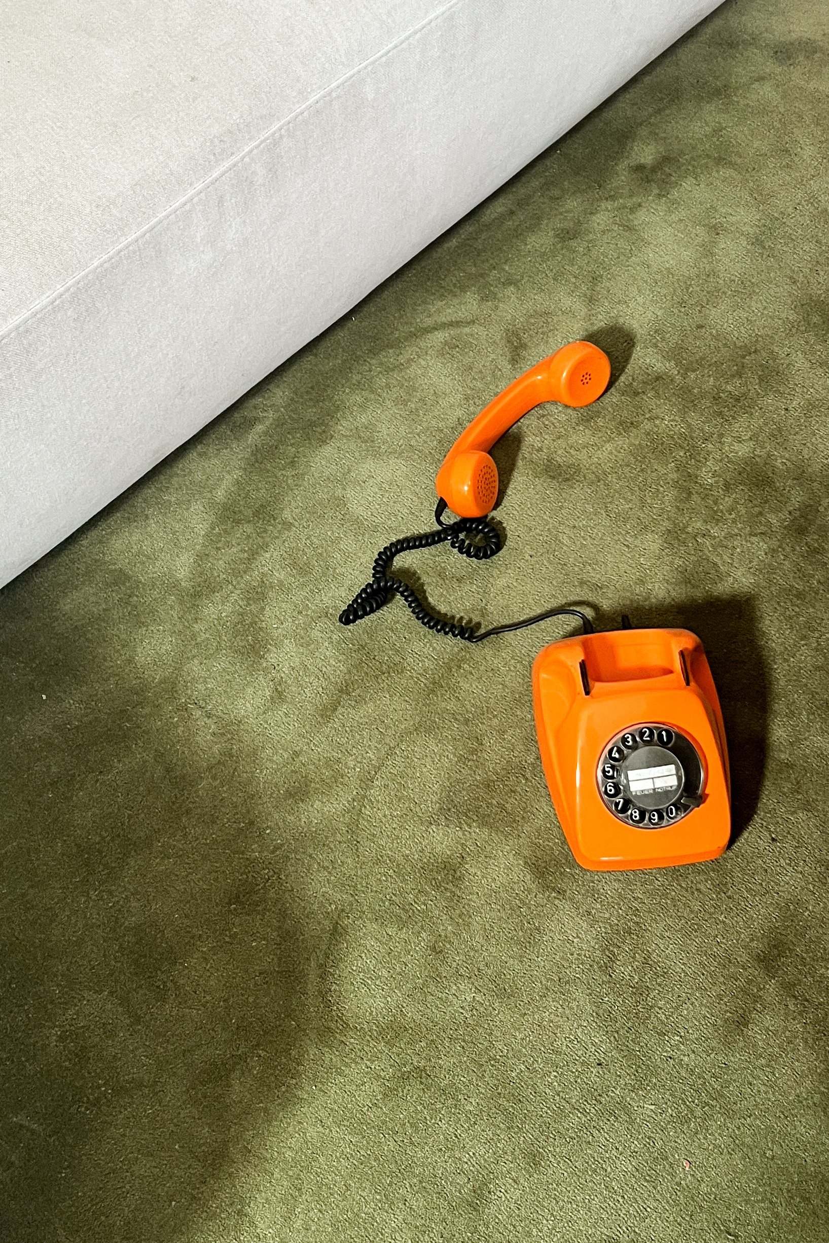 An orange vintage rotary dial telephone resting on green carpet next to a white furniture piece.