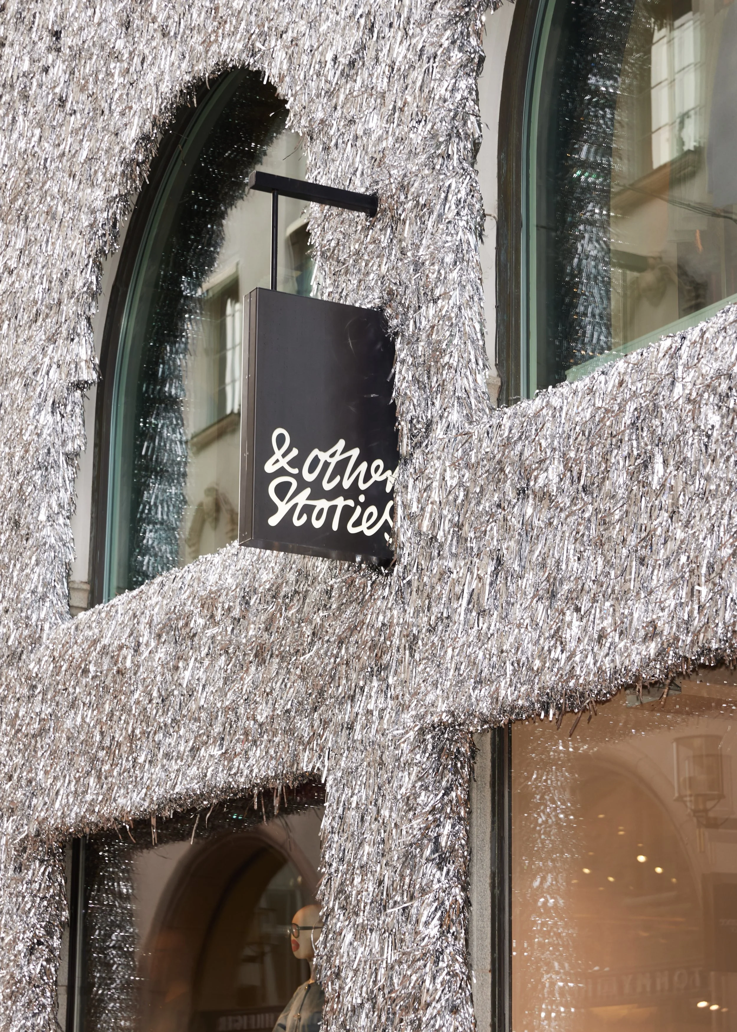 Storefront decorated with silver tinsel and a black sign reading '& other Stories' positioned near a window with a mannequin wearing glasses inside.