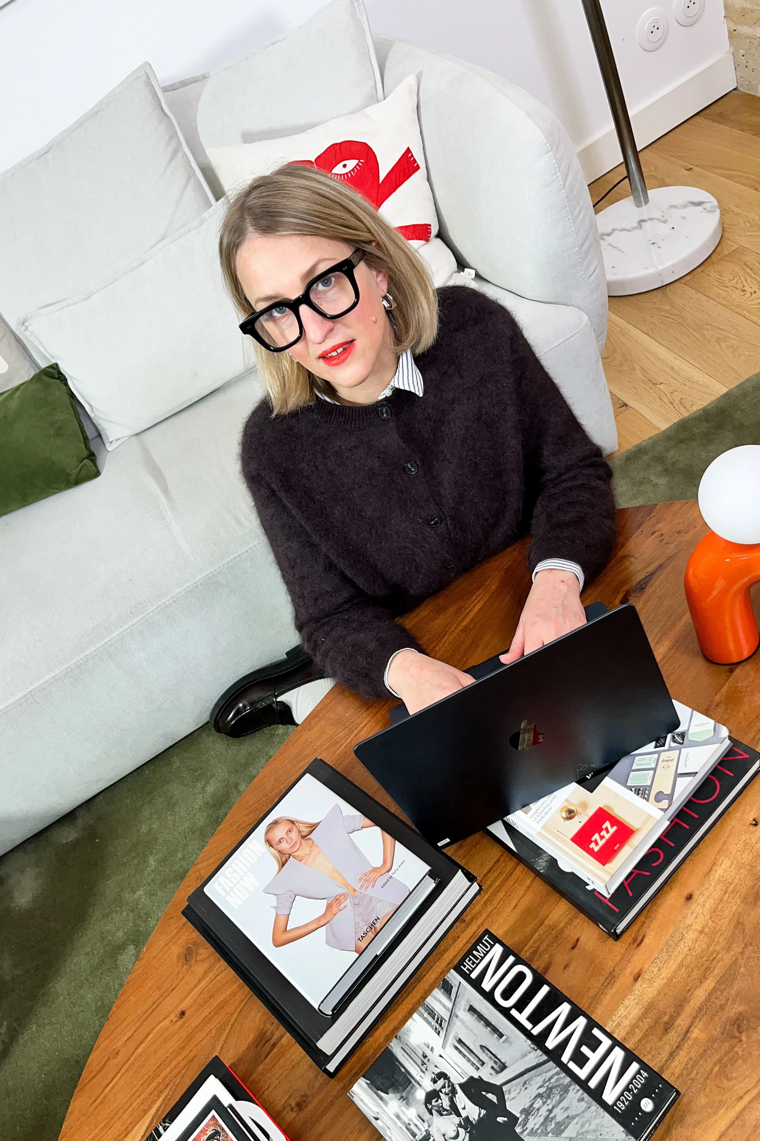 A woman with blonde hair, black glasses, red lipstick, wearing a black sweater over a collared shirt, sitting at a wooden table with a laptop, magazines, and books, in a living room with a white sofa and decorative pillows.