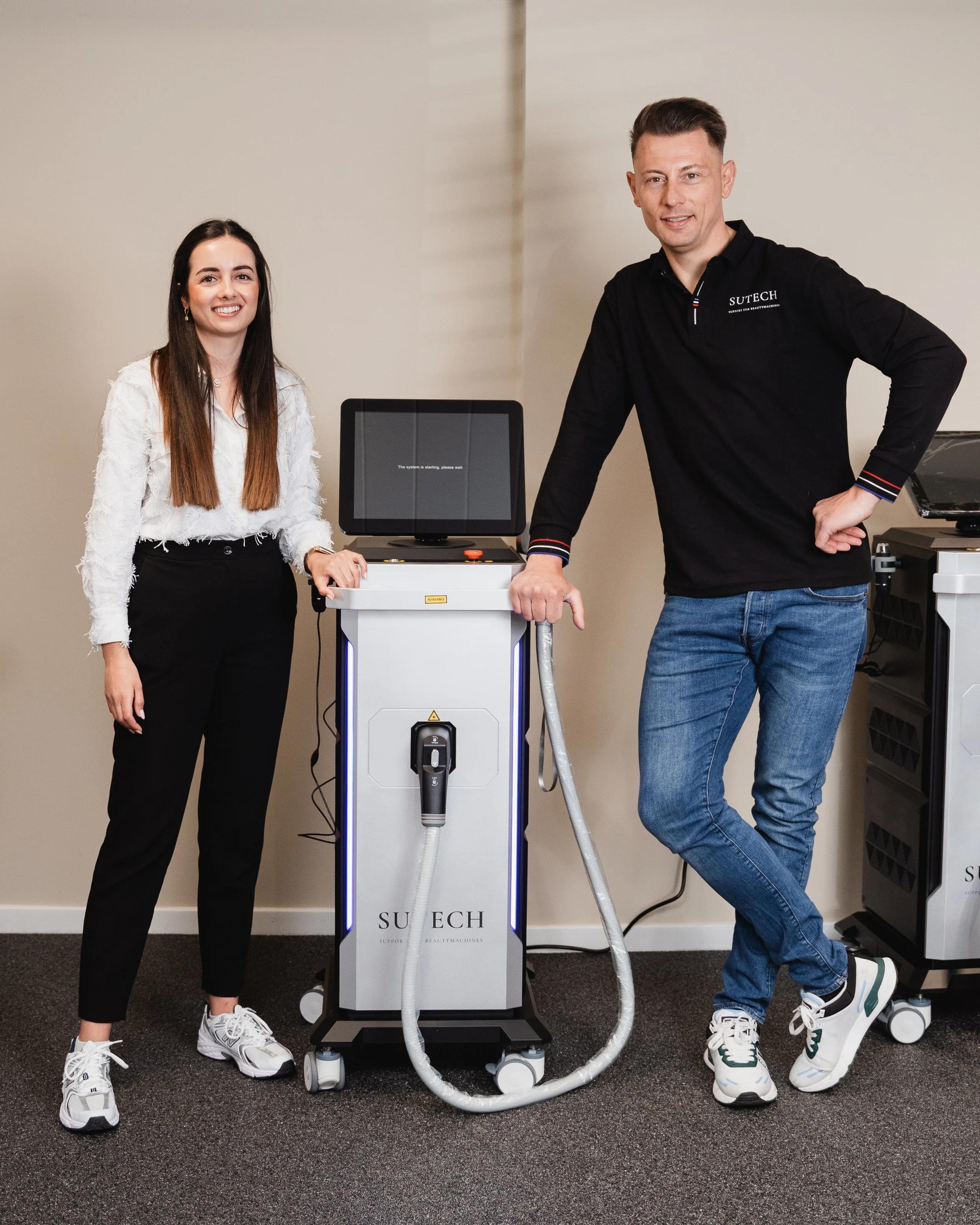 Two people standing next to medical support equipment, a woman on the left wearing a white blouse and black pants, and a man on the right wearing a black long-sleeve shirt and blue jeans. The equipment has a screen displaying a message and the word 'SUTECH' on it.