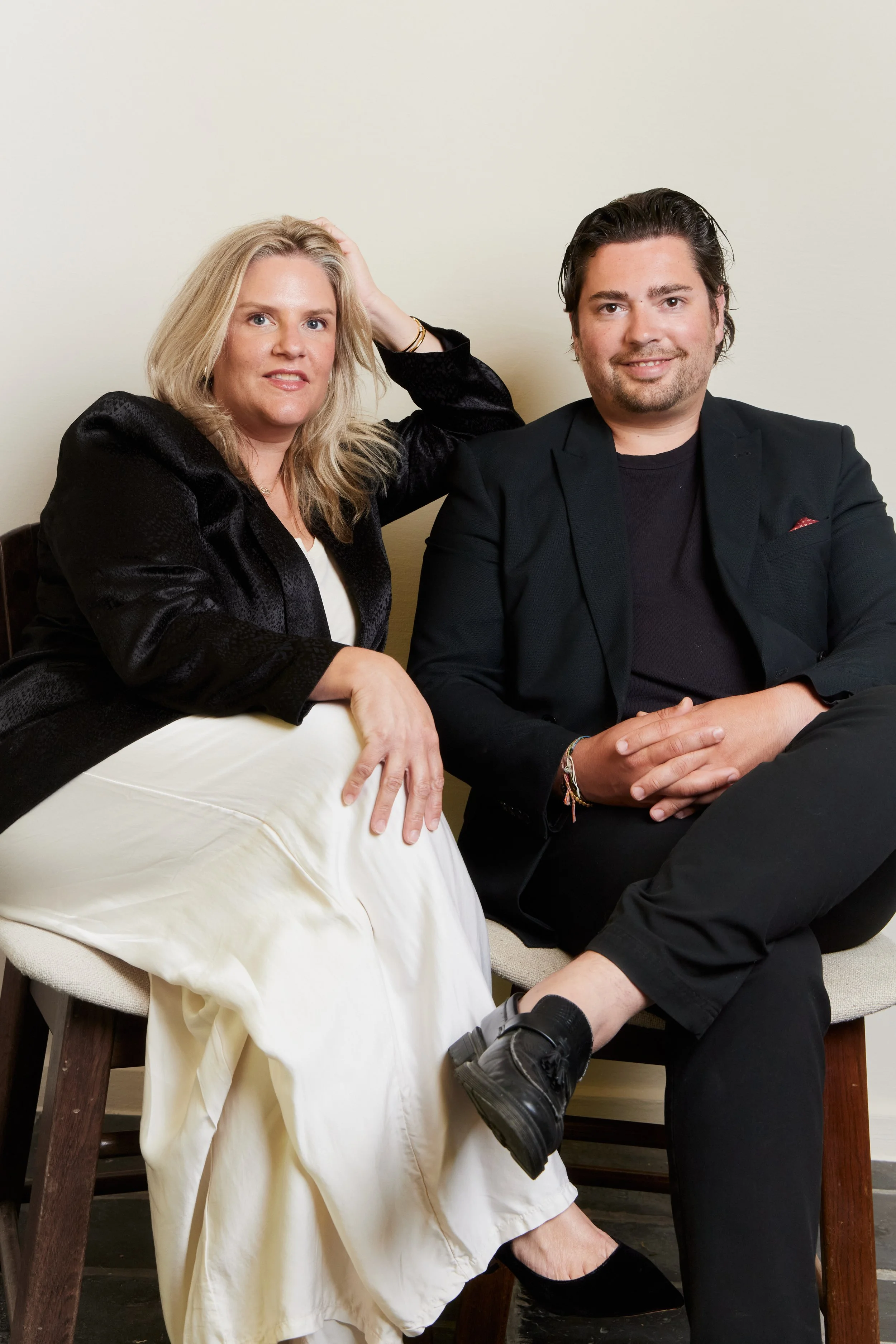 A woman with blonde hair wearing a black blazer and white dress, sitting next to a man with dark hair wearing a black blazer and black pants, sitting on chairs against a plain background.
