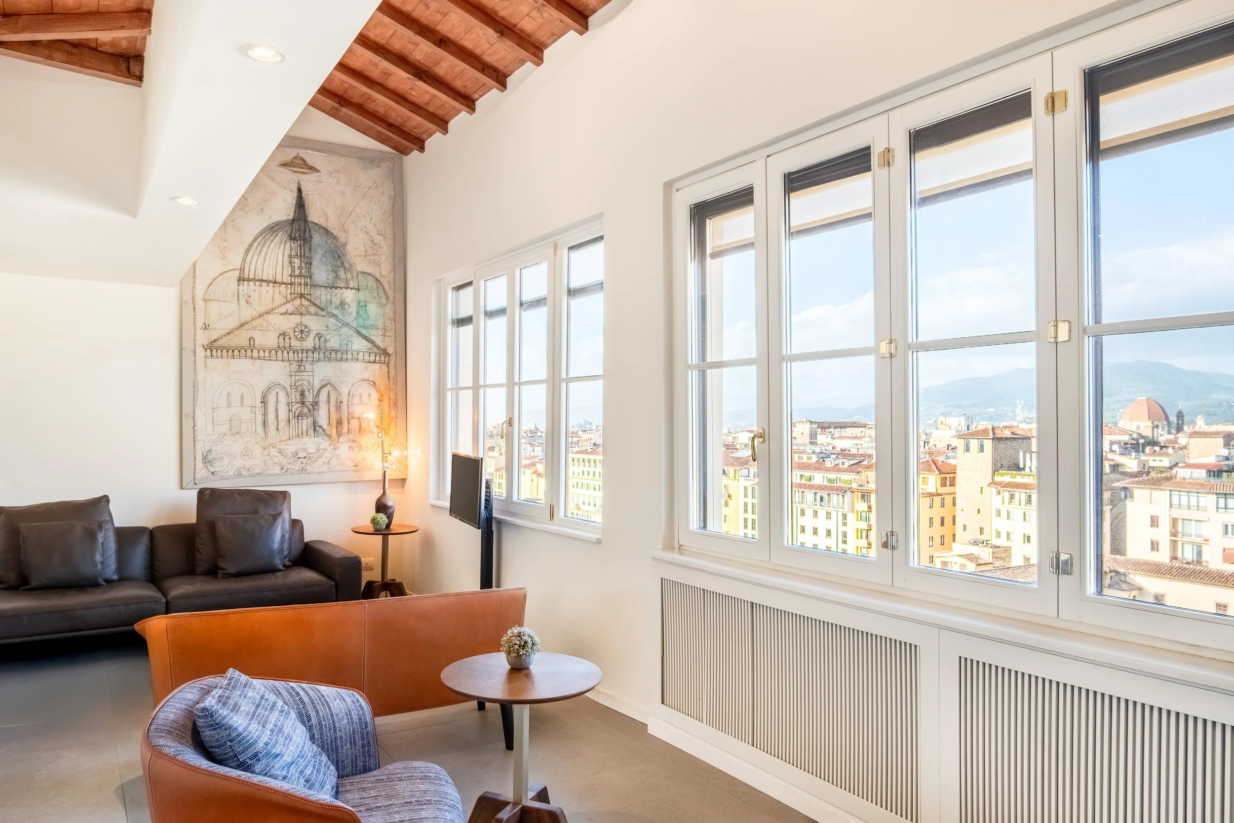 Interno di un soggiorno con grandi finestre che offrono una vista sulla città e le montagne, arredi moderni e un quadro con uno schizzo di una chiesa sul muro.