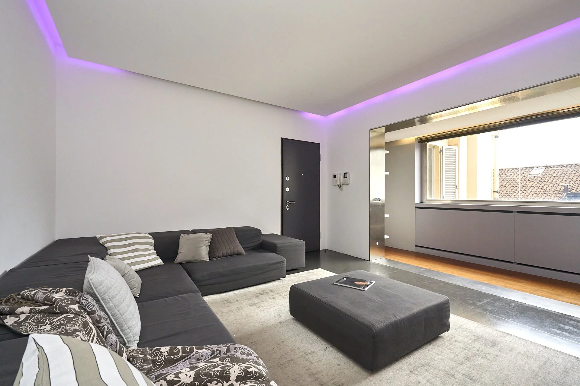 Soggiorno moderno con divano nero, tavolino grande nero, tappeto chiaro, pareti bianche, con illuminazione a LED viola nel soffitto, finestra con vista sui tetti delle case.