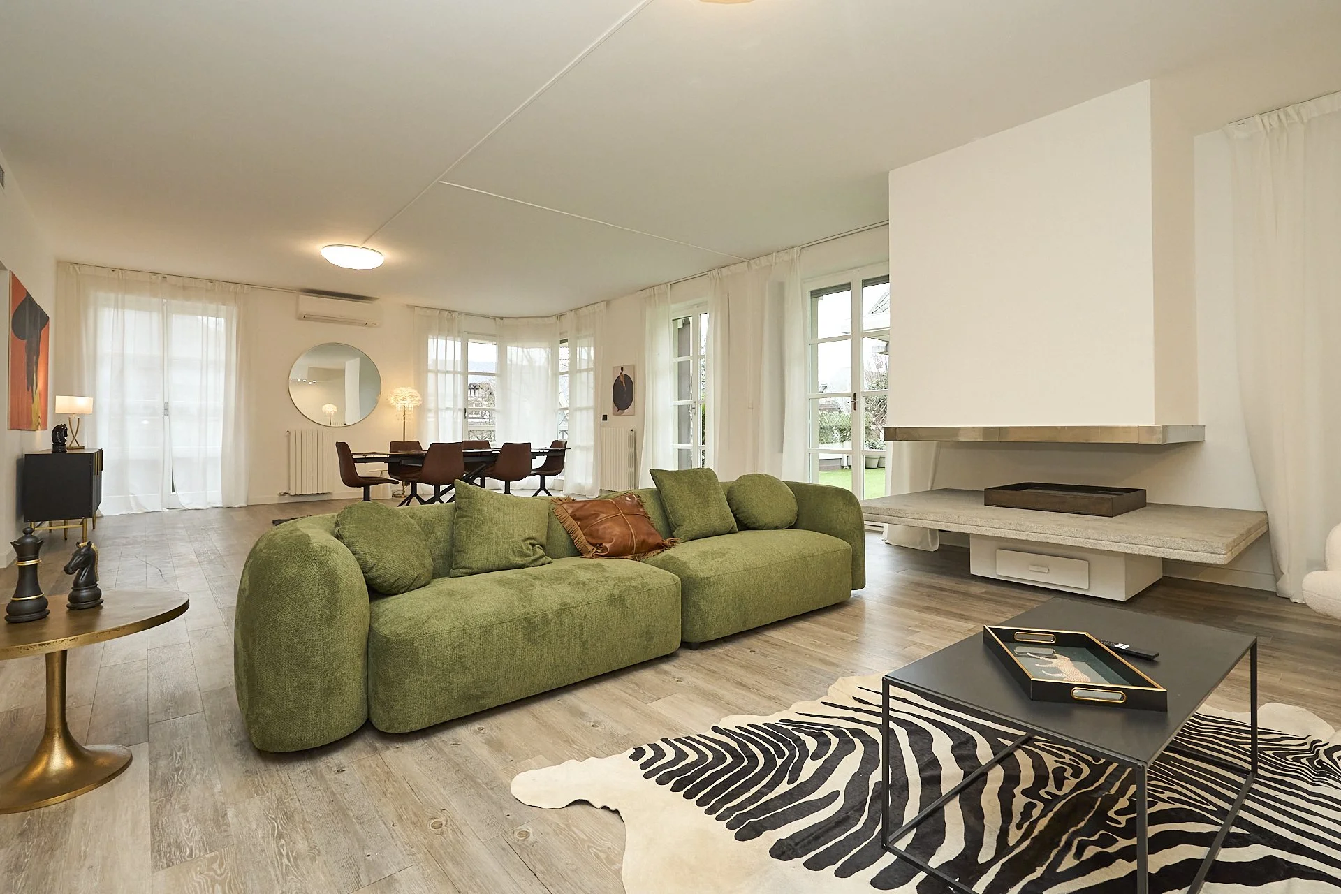 Soggiorno moderno con divano verde, tavolino nero, tappeto zebra, pareti bianche, grandi finestre con tende bianche, sala da pranzo con tavolo e sedie marroni, decorazioni moderne.