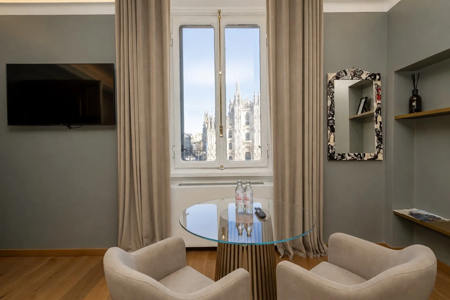 Interno di una stanza con una finestra che mostra il Duomo di Milano. Al centro una tavola rotonda con due bottiglie d'acqua, un telecomando e due sedie beige.