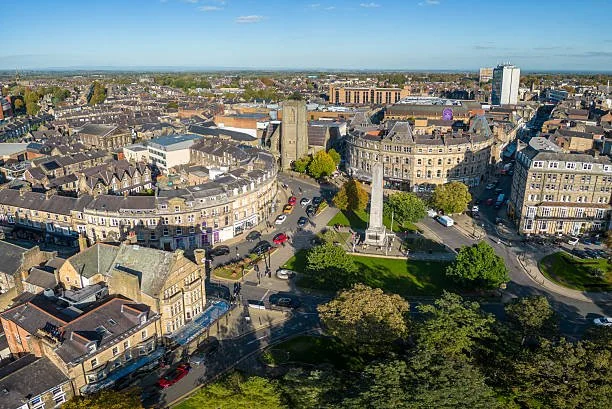 Aerial view of a city square with historic buildings, a tall monument, and green park area surrounded by trees and streets, under a clear blue sky.