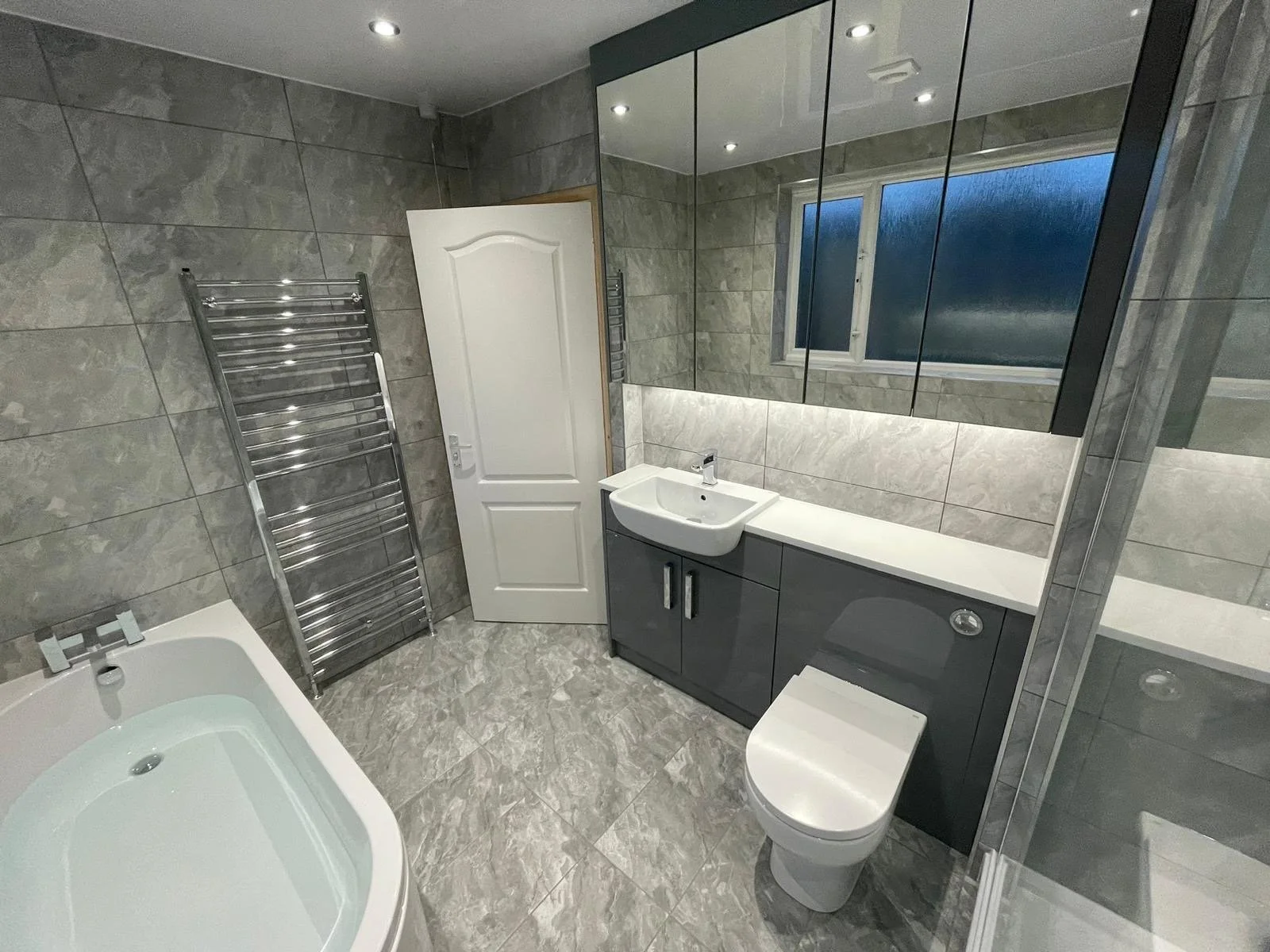 Modern bathroom with gray tiled walls and floor, a white bathtub, a gray vanity with a sink, a large mirror, a window, and a white door.