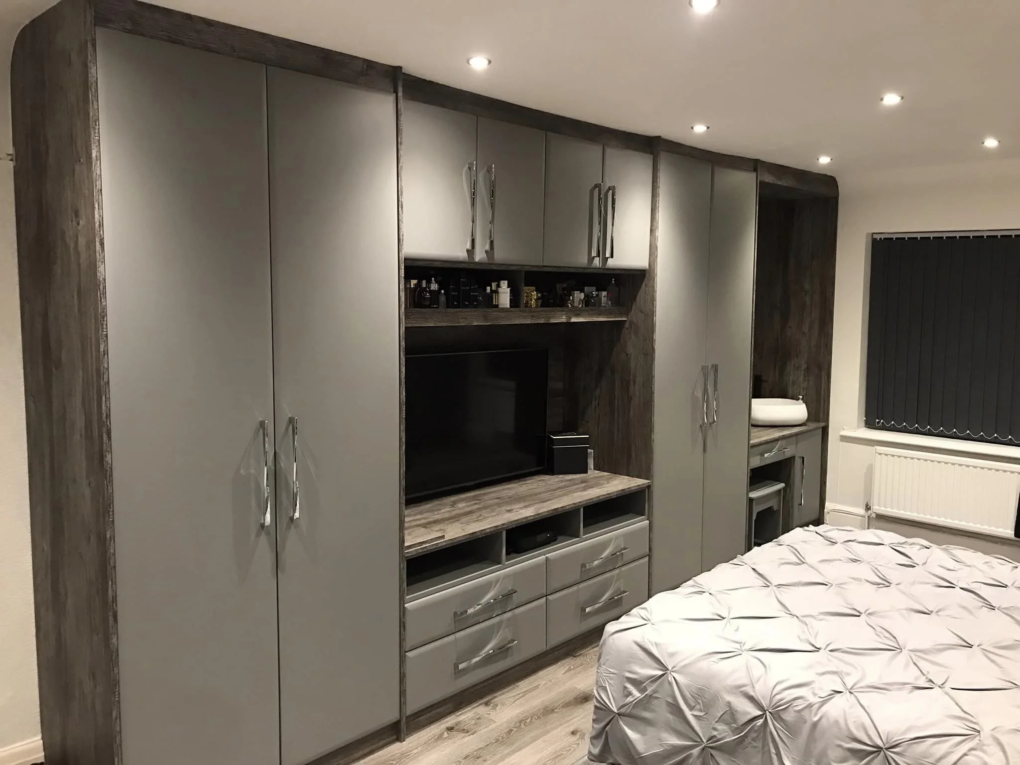Bedroom with built-in gray and wood storage unit, flat-screen TV, and a bed with a tufted white bedspread. Window with black blinds and a radiator below.