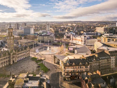 Aerial view of a city square with historic and modern buildings, a clock tower, and a fountain, under a partly cloudy sky.