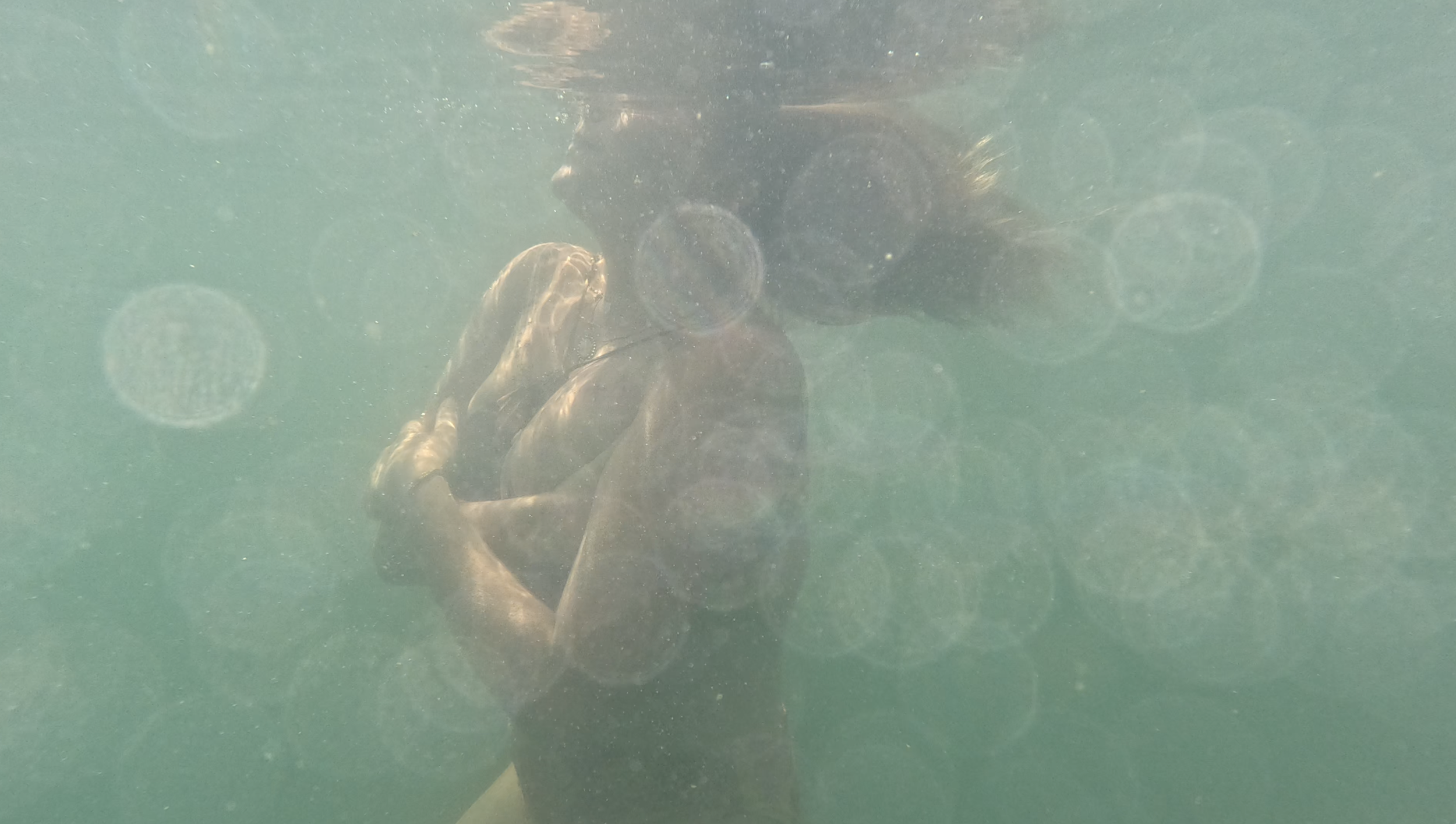 Eine Person, die unter Wasser sitzt, umarmt sich selbst und hat die Haare offen. Im Hintergrund sind Wasserblasen und Lichtreflexionen.