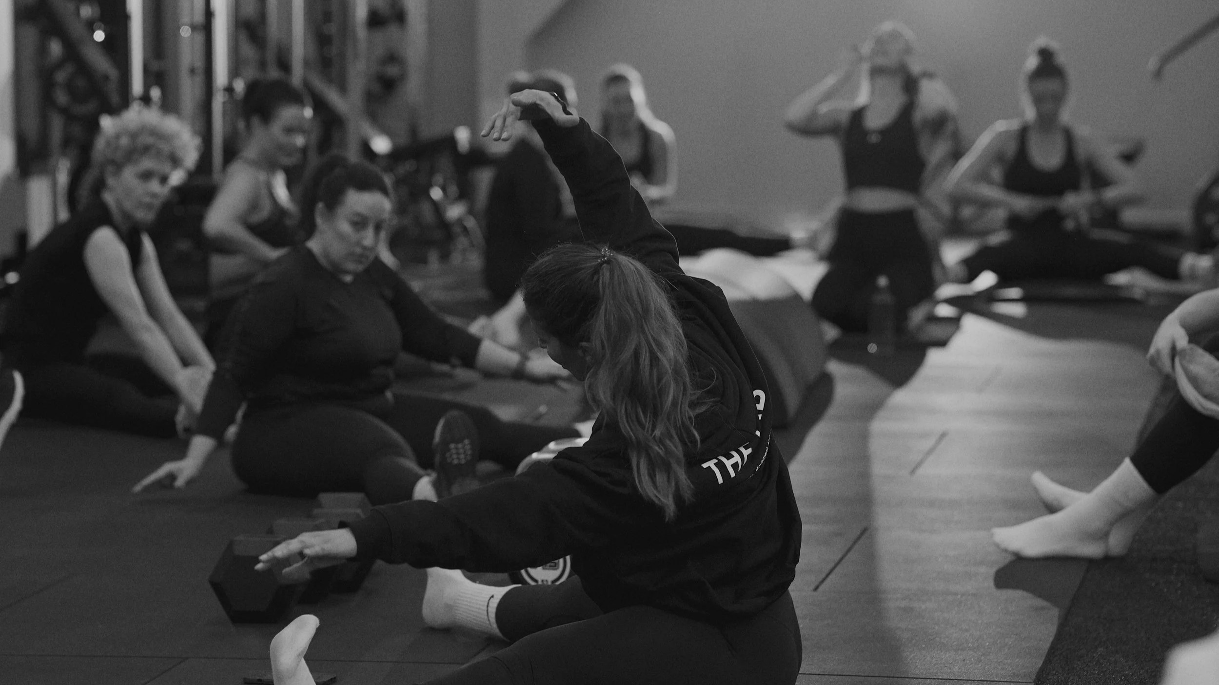 Women participating in a seated yoga or stretching class on mats in a gym, with one woman in front demonstrating a stretch.