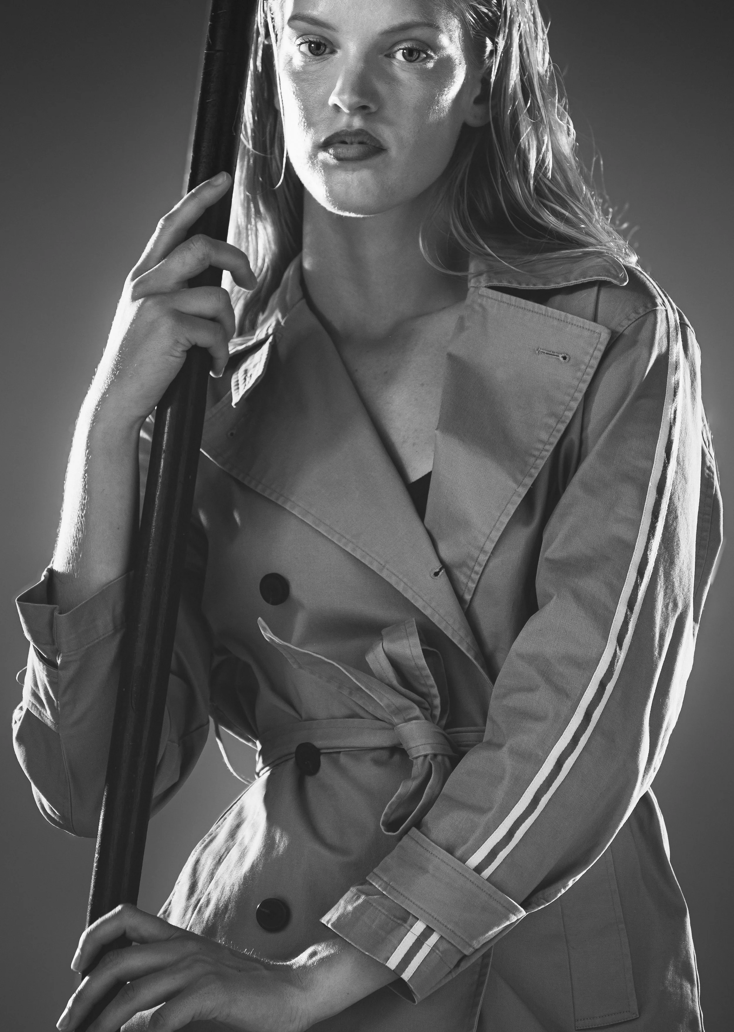 Une femme portant un trench beige, tenant un parapluie, photographiée en noir et blanc.