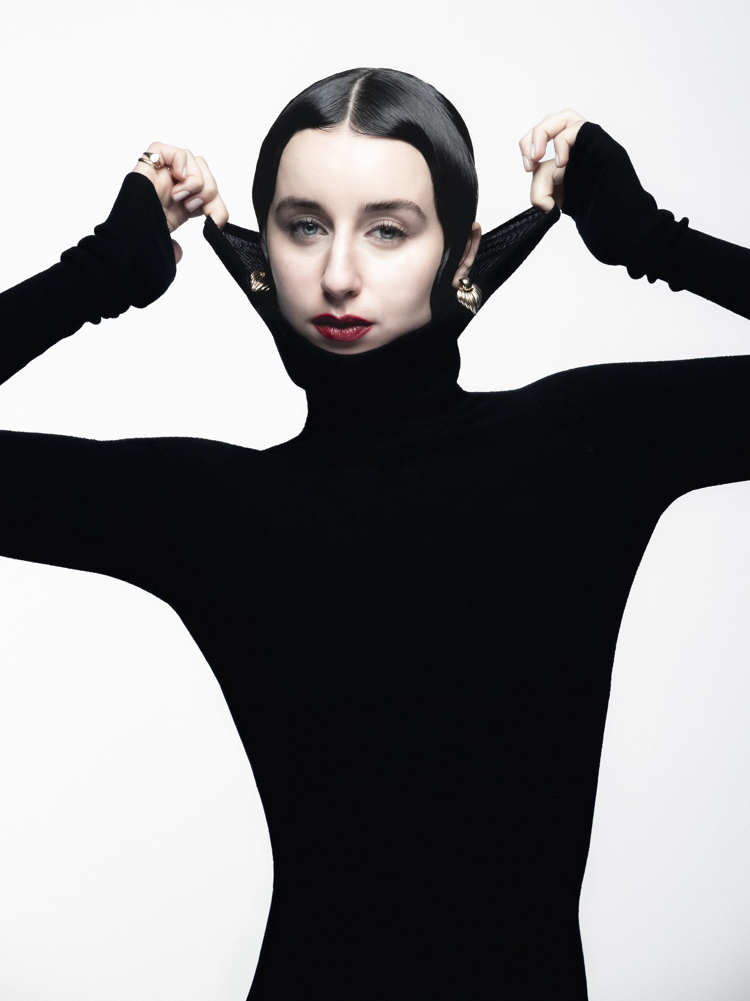 Une femme avec cheveux noirs, portant un turtleneck noir, tire sur le col de son vêtement, fond blanc, maquillage rouge à lèvres.