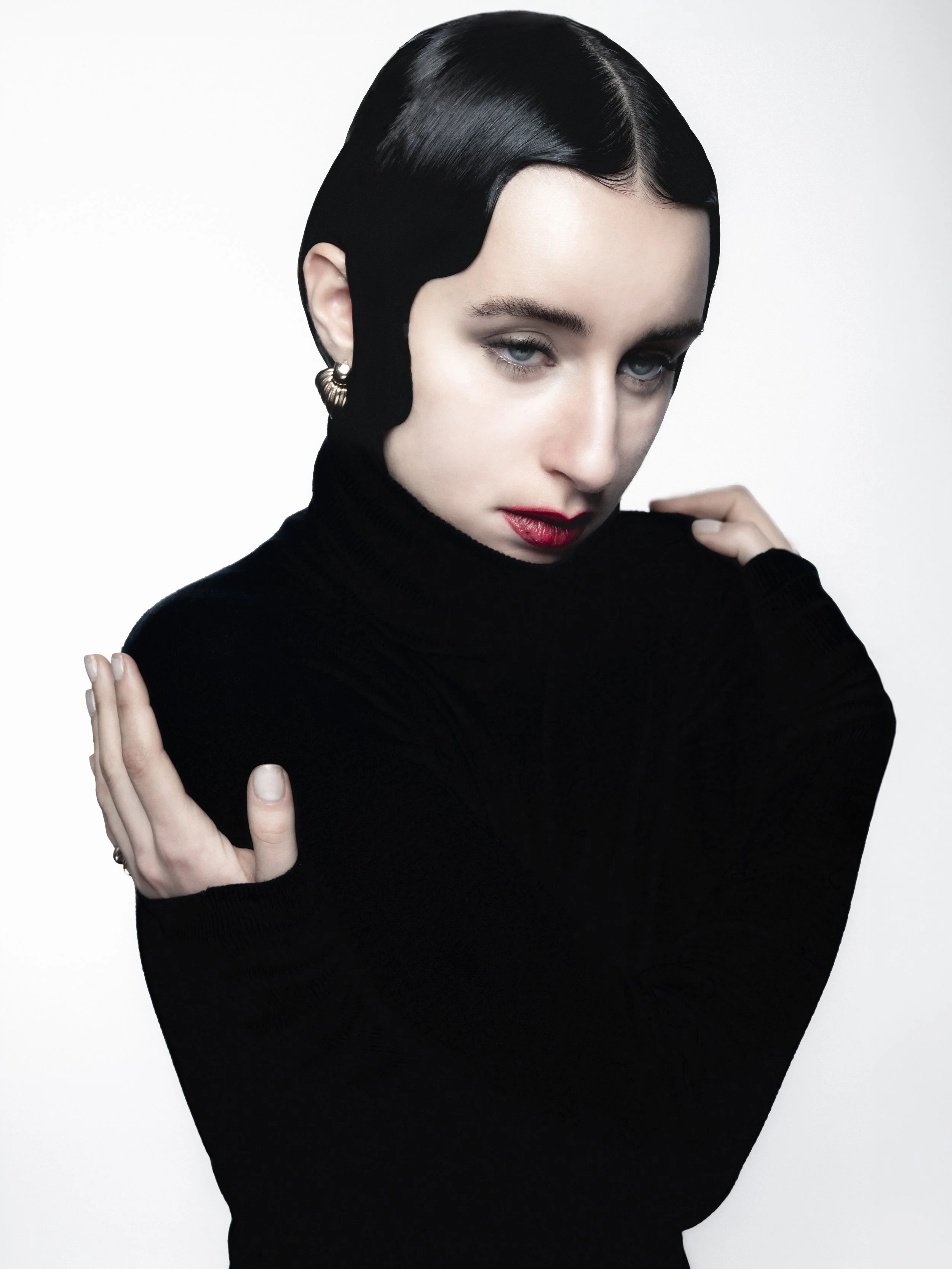 Portrait d'une femme avec des cheveux coiffés en ligne de côté, portant un pull noir à col roulé, avec des boucles d'oreilles en or, maquillage rouge à lèvres et expression intense.