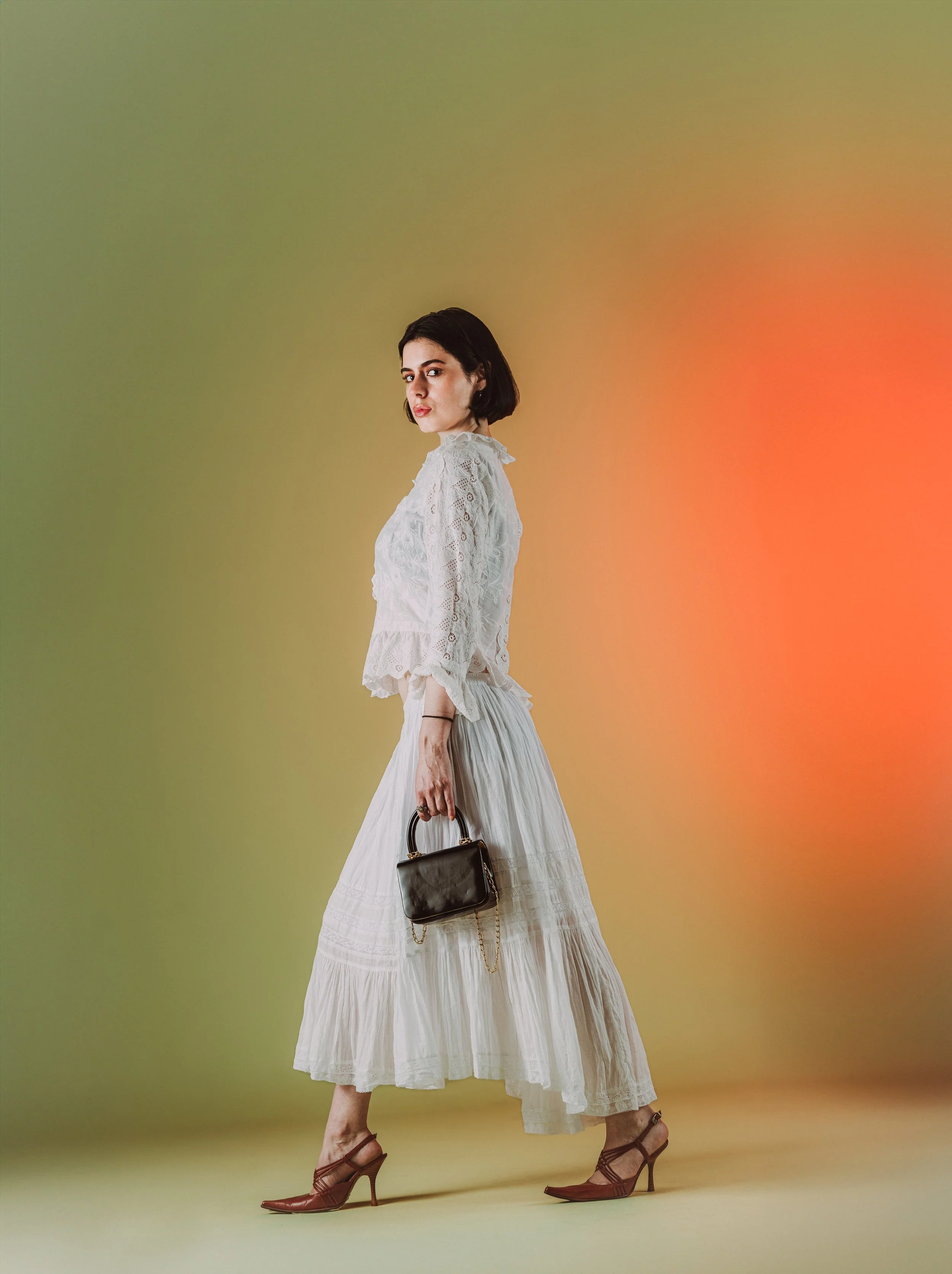 Femme portant une robe longue blanche, portant un sac à main noir, sur un fond coloré dégradé.