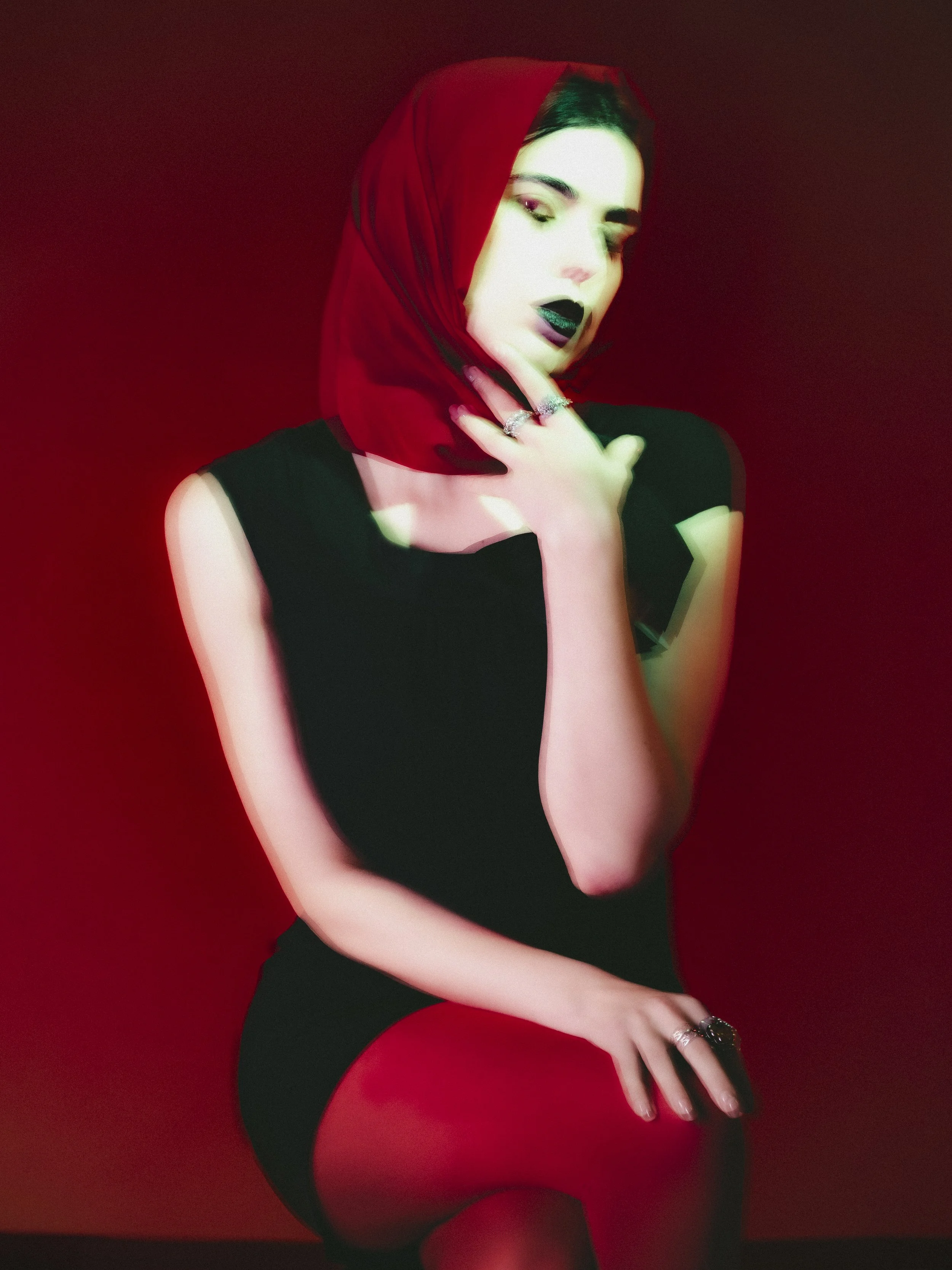 Une femme portant un turban rouge, vêtue d'une robe noire, avec des lèvres foncées, posant contre un fond rouge.