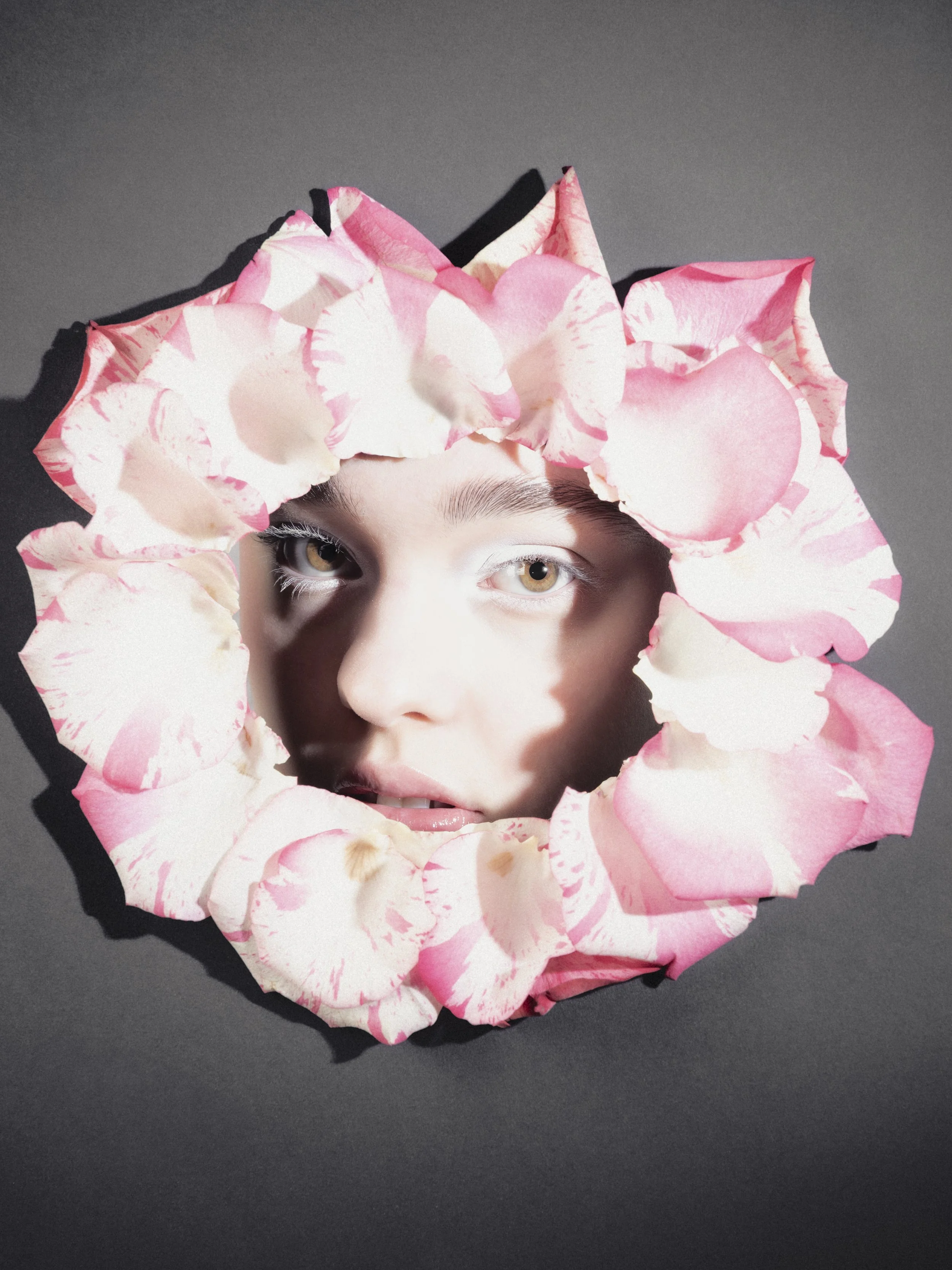 Un visage humain regardant vers l'objectif, encadré par des pétales de fleurs roses et blanches disposés en cercle.