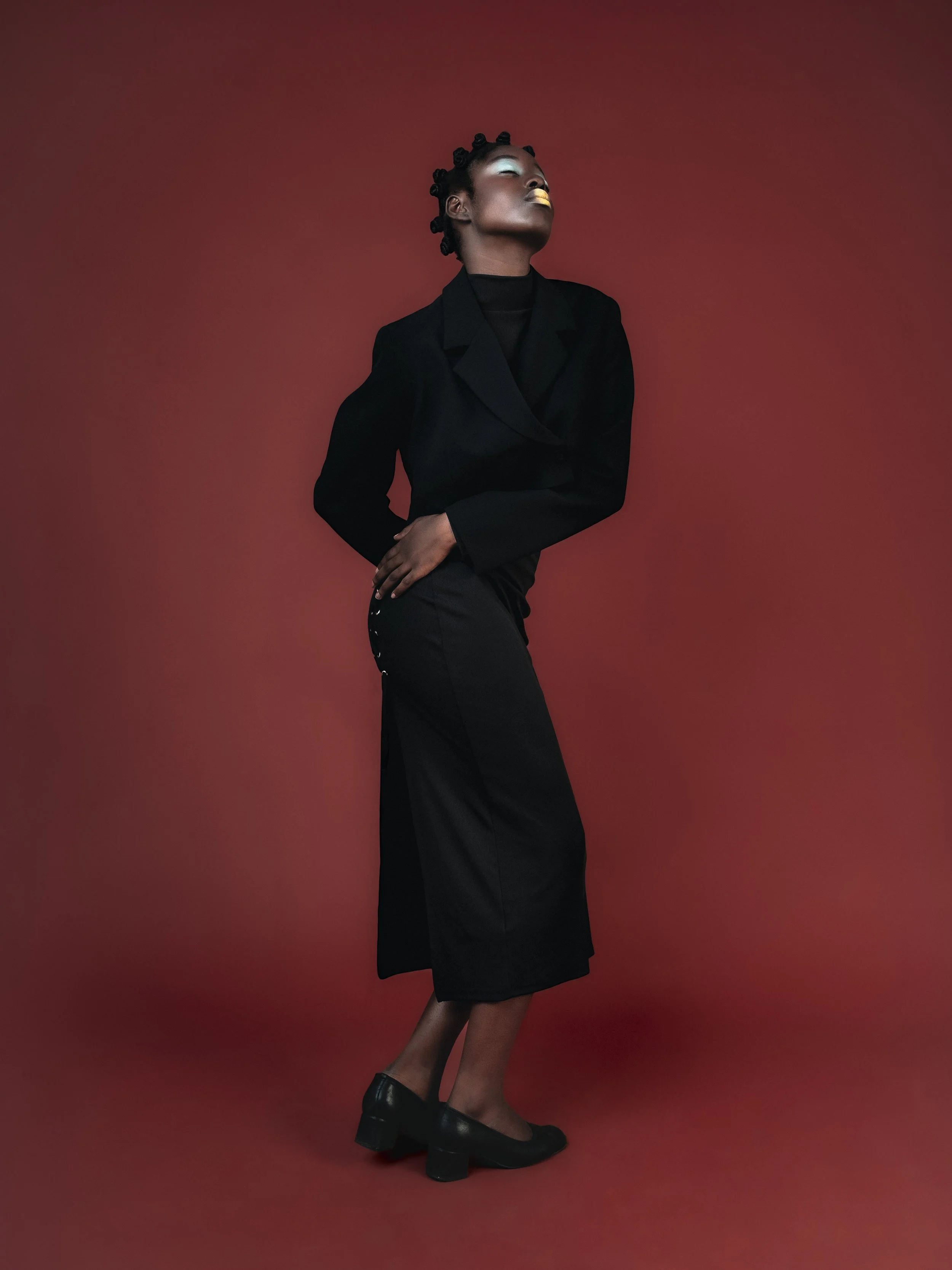 Une femme noire pose contre un fond rouge, portant un costume noir et des chaussures noires, avec une coiffure en petits chignons et un maquillage sophistiqué.