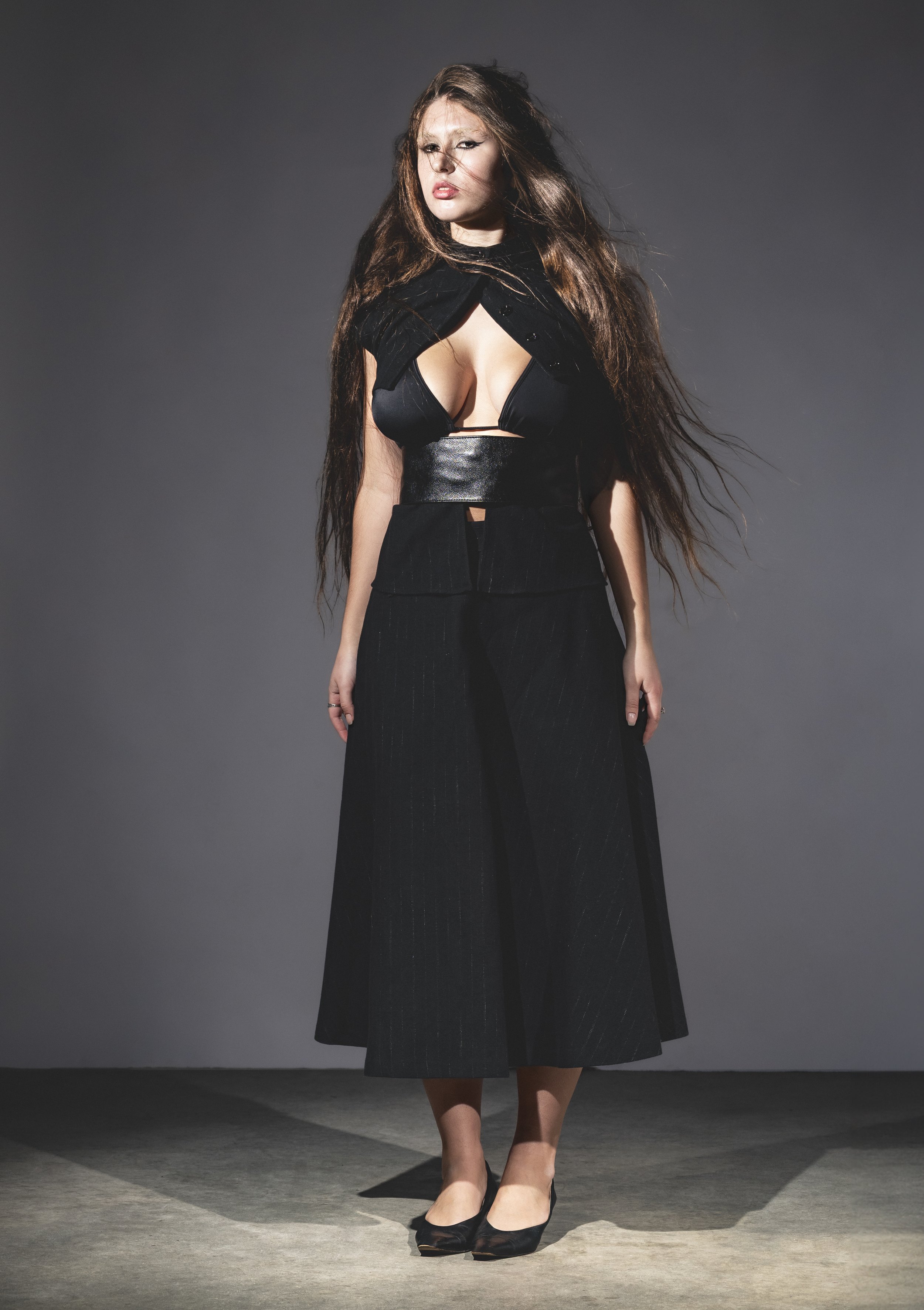 Une femme portant une tenue noire avec du cuir et un décolleté profond, posant sur un fond gris, avec des cheveux longs et bouclés.