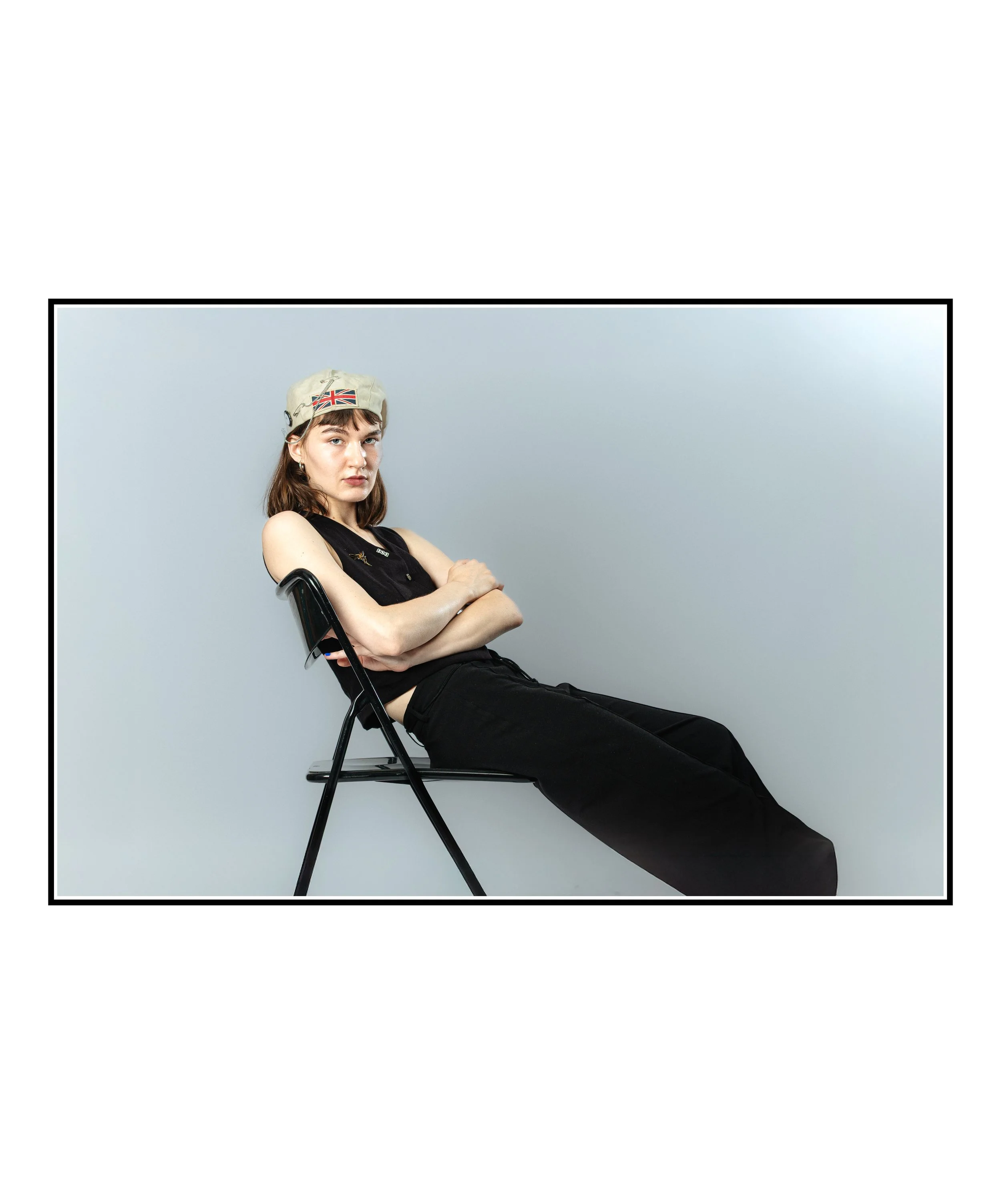 Jeune femme assise sur une chaise, portant un t-shirt noir, un pantalon noir, un béret avec le drapeau britannique, croisant les bras, regardant la caméra avec une expression neutre, fond gris clair.