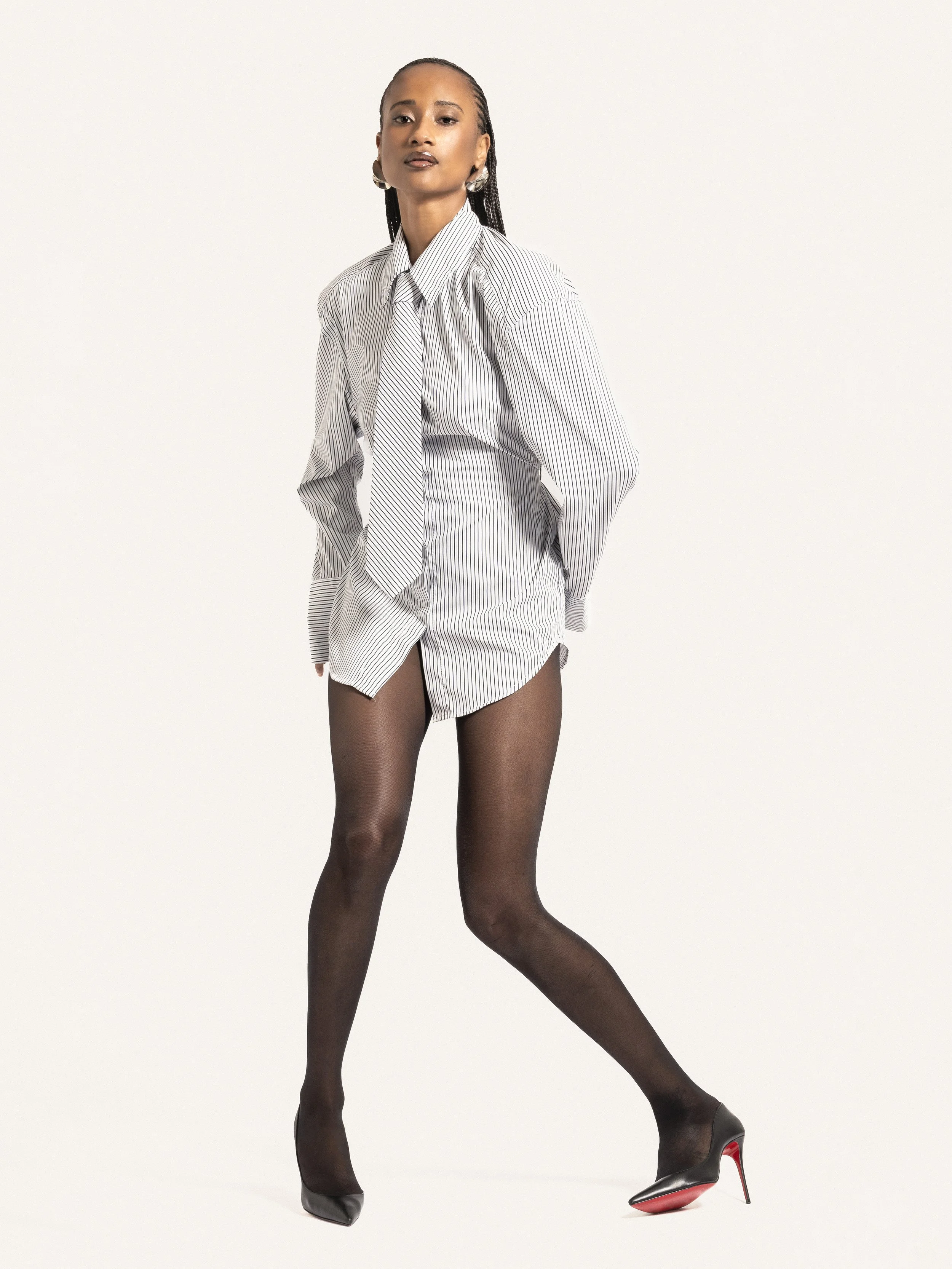 Femme portant une chemise blanche à rayures, des collants noirs et des talons noirs avec semelles rouges, debout contre un fond blanc.