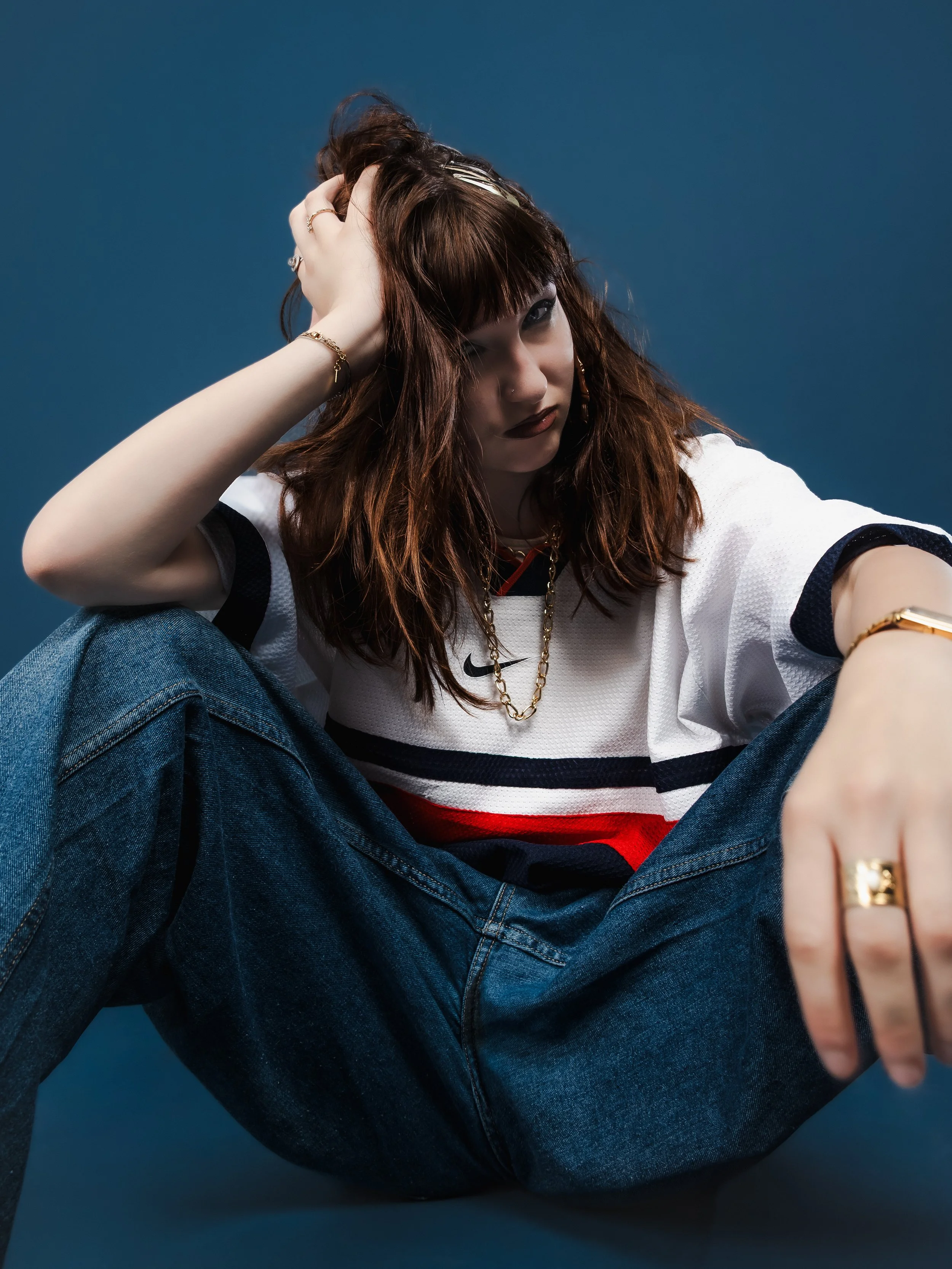 Jeune femme aux cheveux bruns, vêtements décontractés, posant contre un fond bleu, assise avec jambes croisées, portant des bijoux en or, et regardant la caméra