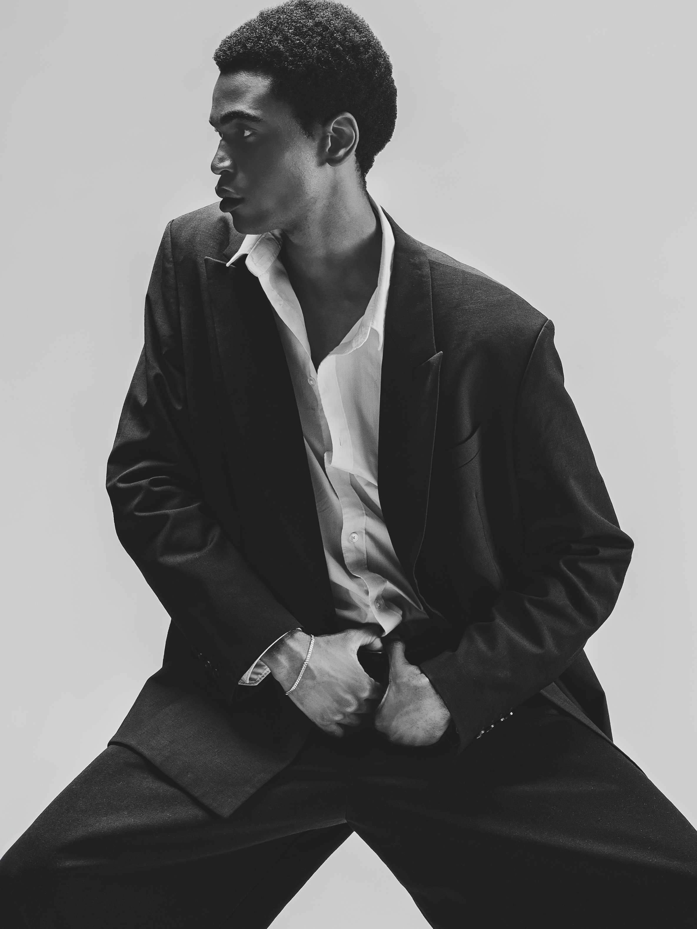 Un homme en costume noir, chemise blanche, pose dynamique en noir et blanc, les jambes écartées et les mains à la taille, regardant de côté.