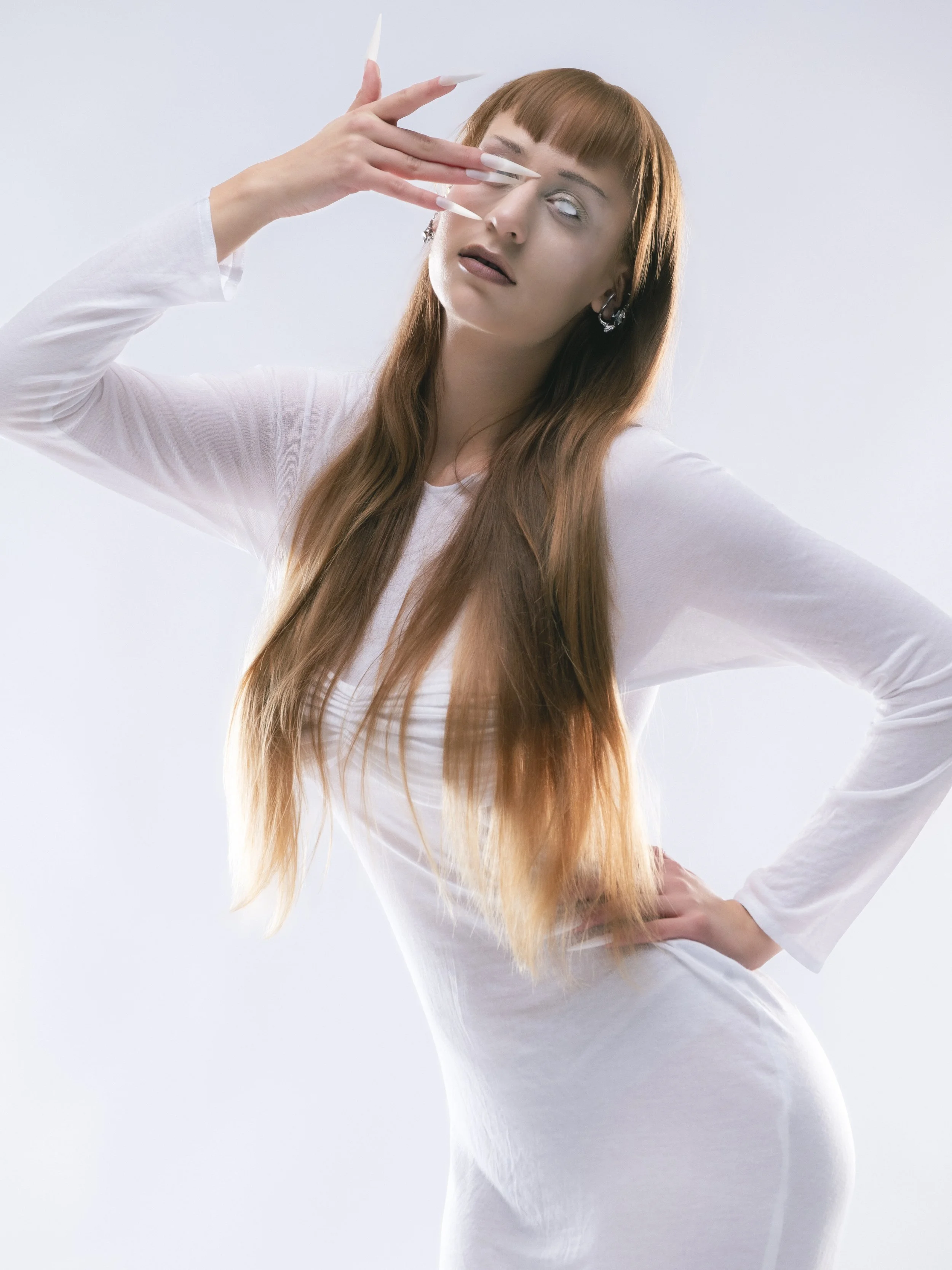 Femme aux longs cheveux marron clair et aux yeux blancs, portant un vêtement blanc, pose avec une main sur la taille et l'autre près du visage, dans un fond blanc