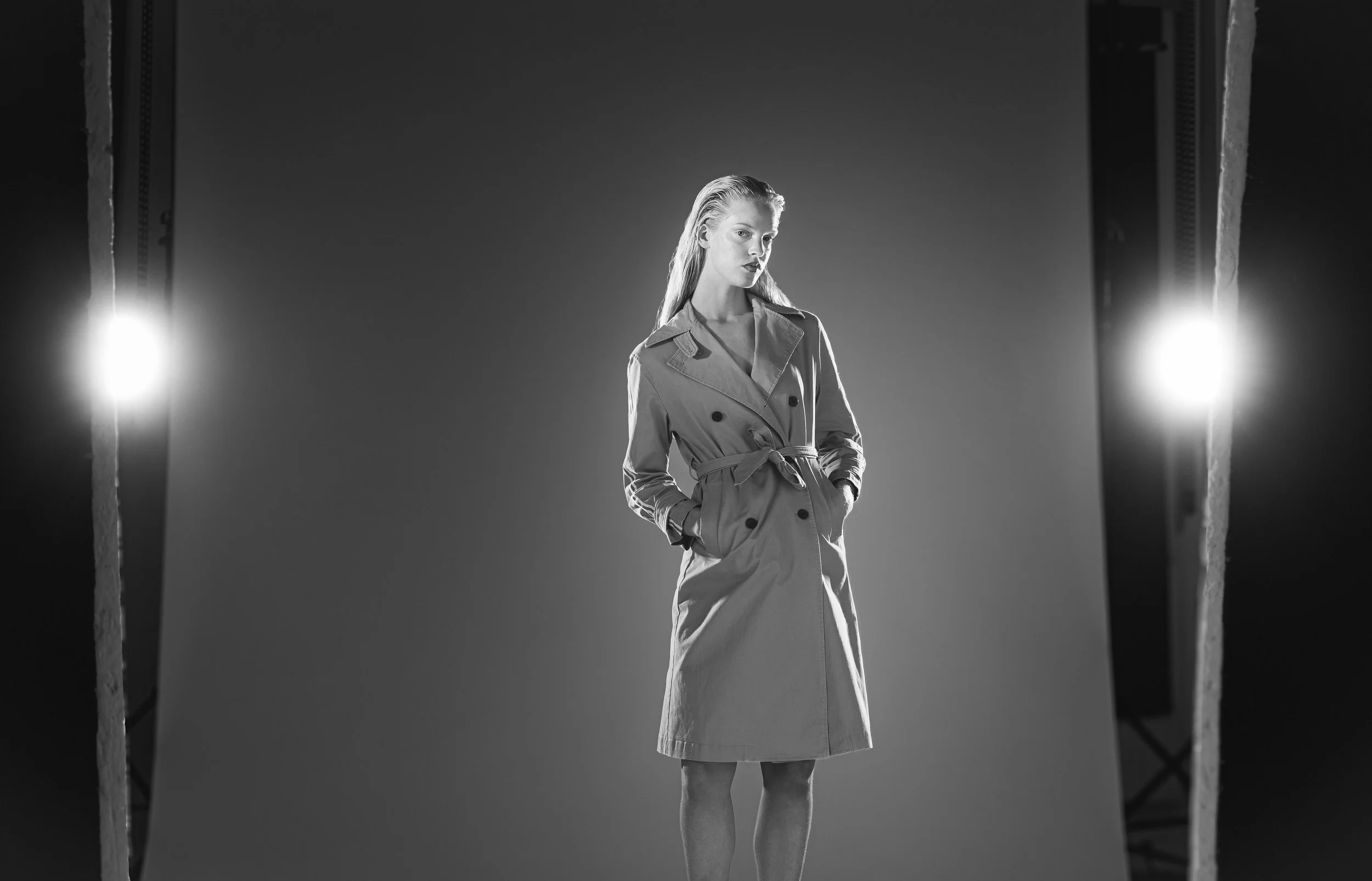 Une femme portant un trench coat, se tenant devant un fond uni, éclairée par deux sources de lumière de chaque côté.