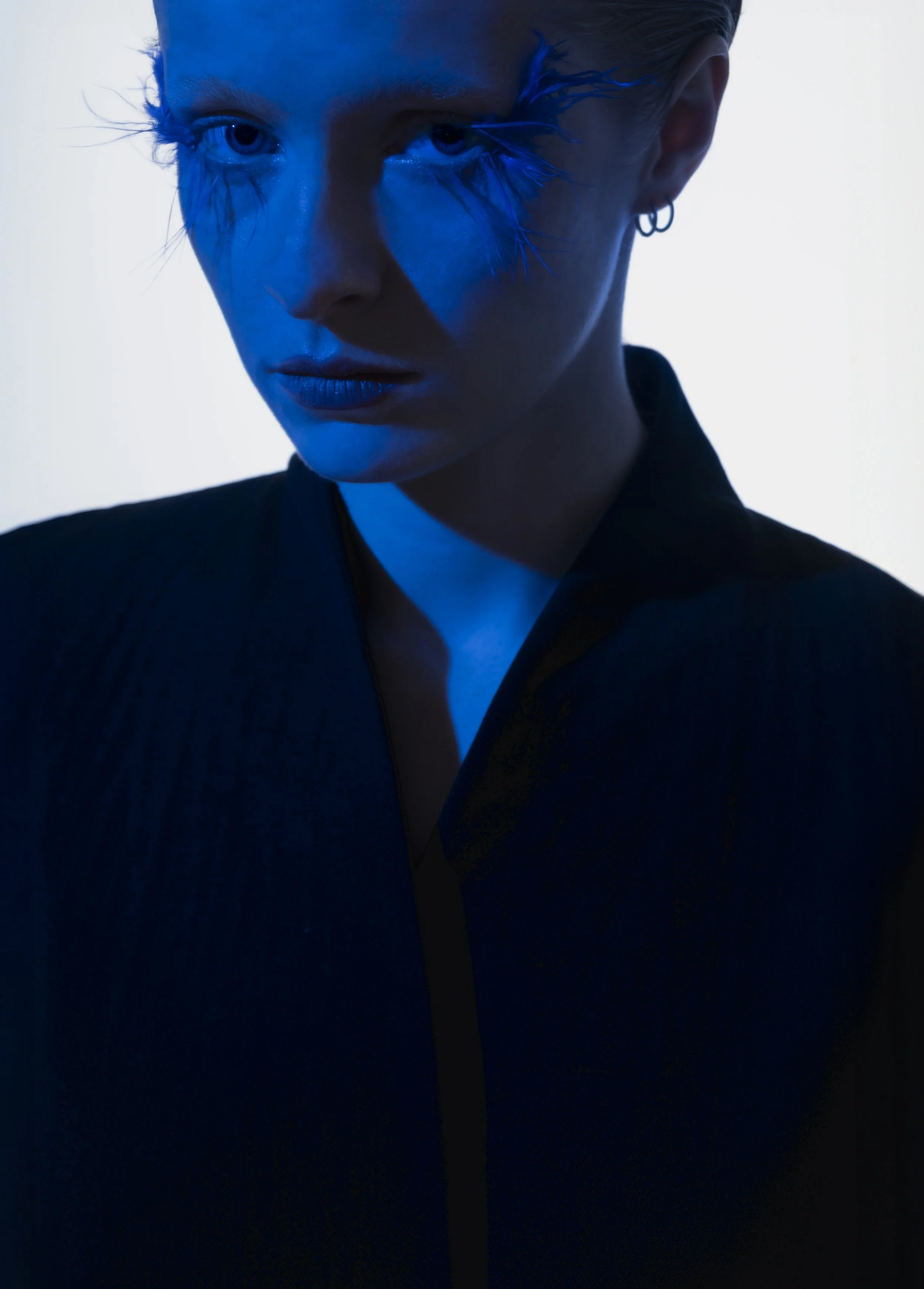 Portrait d'une femme avec maquillage bleu artistique et boucles d'oreilles argentées, portant une veste noire, avec un éclairage froid et un fond clair.