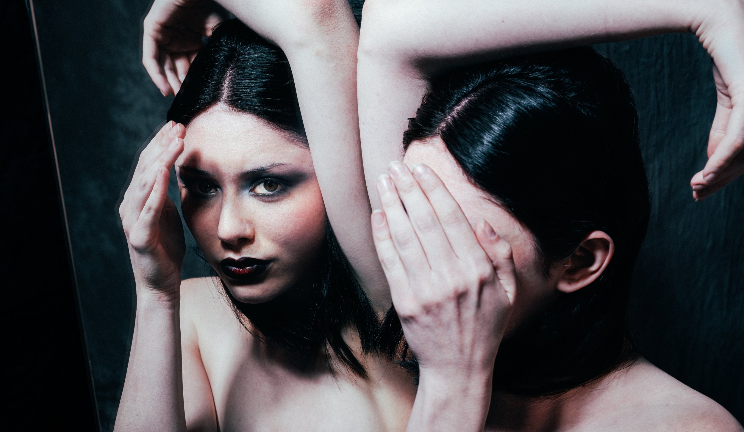 Une femme avec des cheveux noirs et maquillage sombre regarde dans un miroir, avec une main posée sur sa tête et une autre sur son visage.