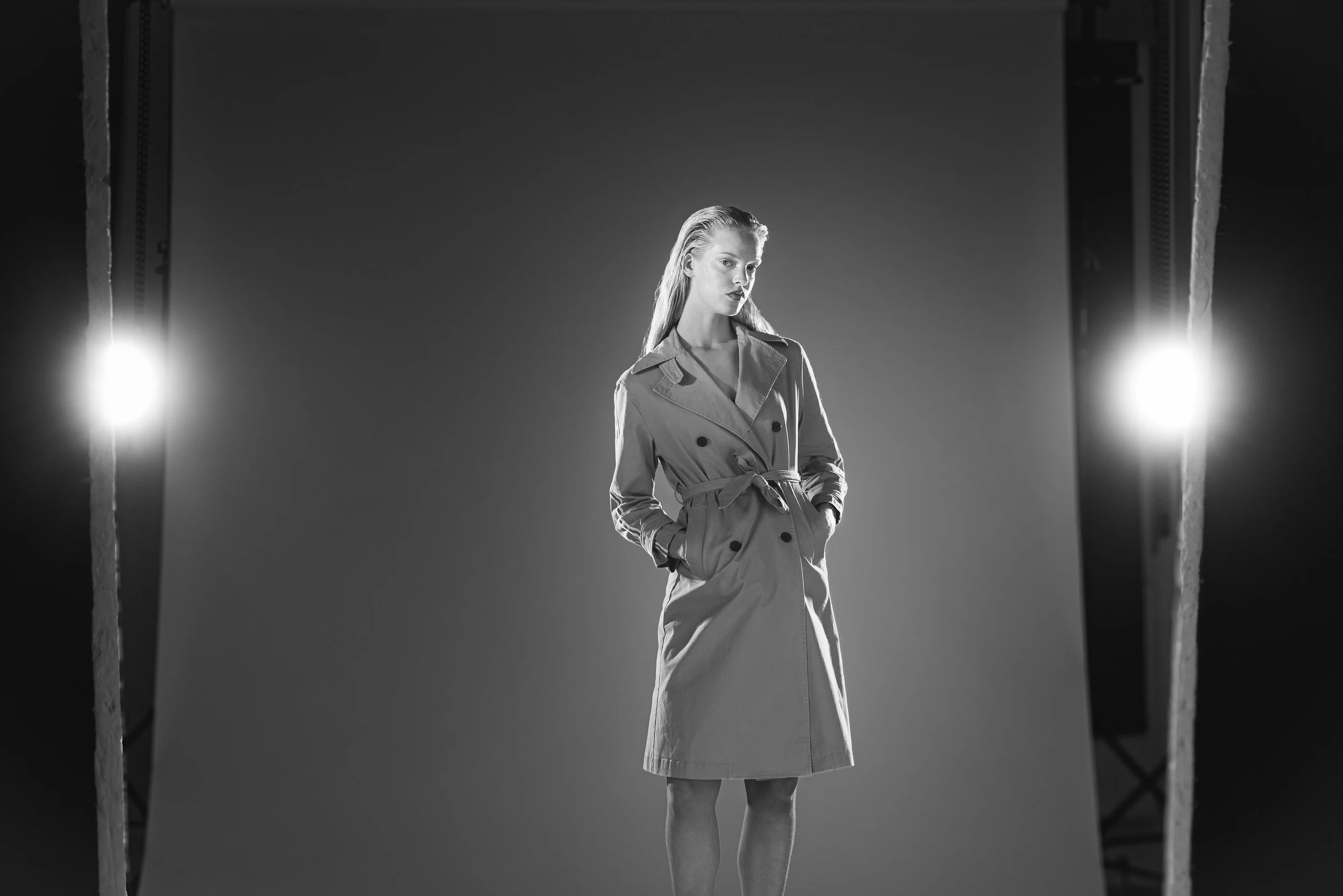 Femme portant un trench beige, debout dans un studio photo avec éclairage brillant de chaque côté.
