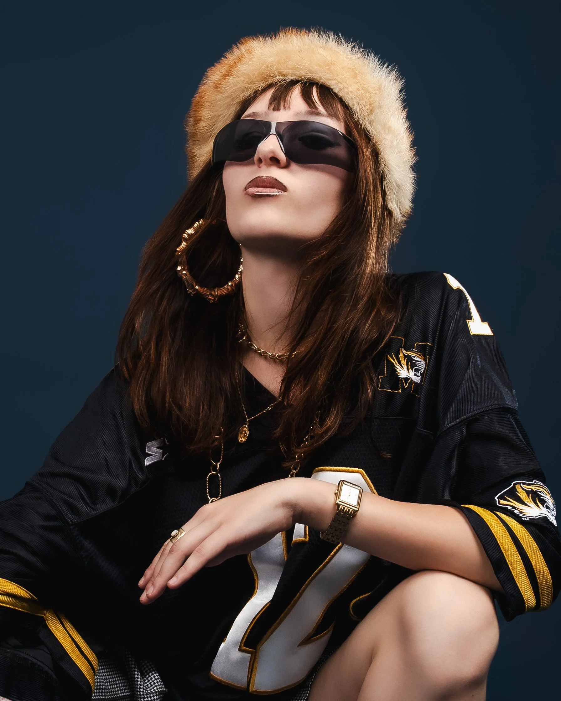 Femme portant un chapeau en fourrure, des lunettes de soleil noires, une veste de football américain noire avec des détails dorés, des boucles d'oreilles en métal argenté, des colliers dorés, une montre en or, et posant avec confiance