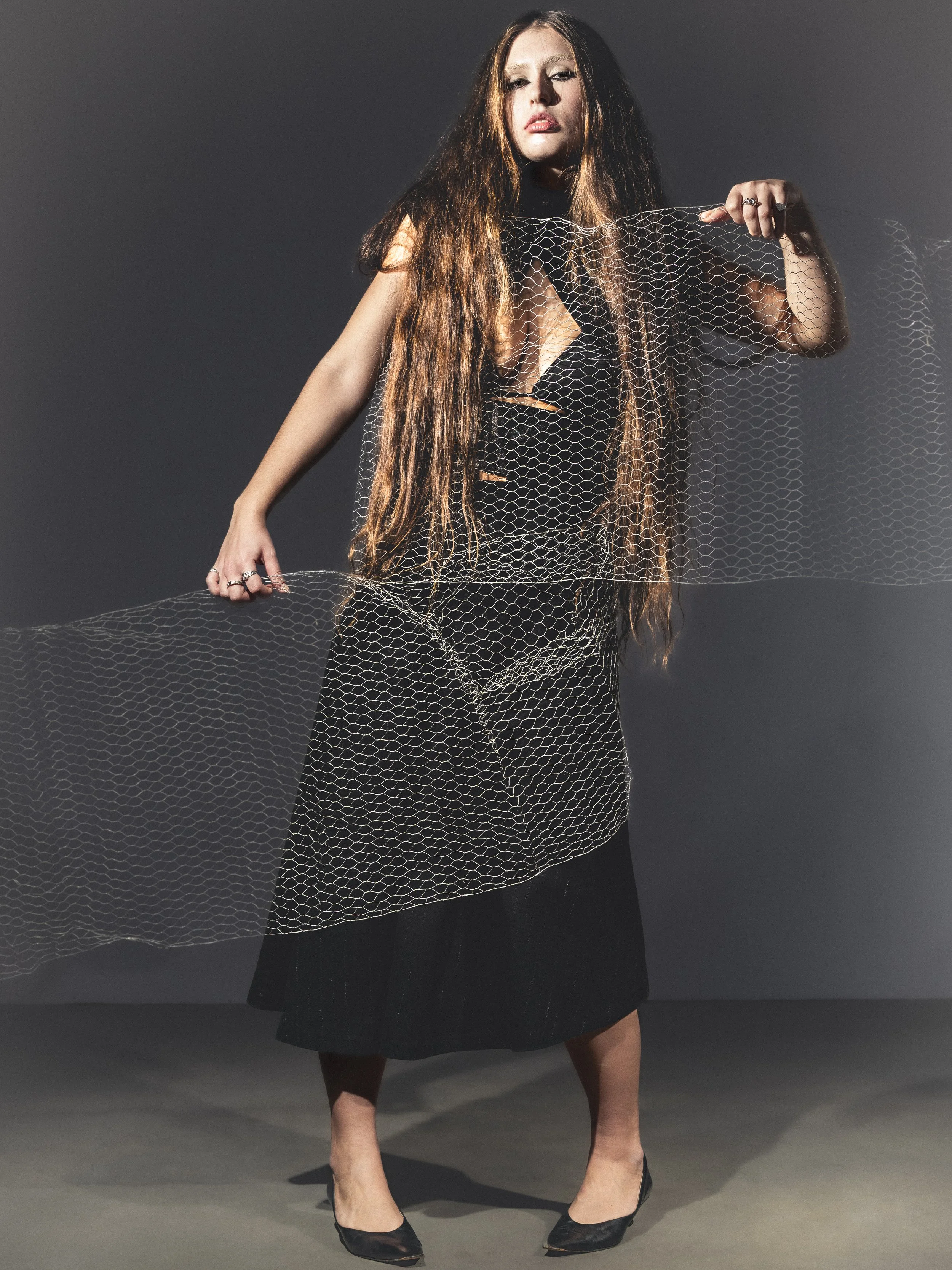 Une femme avec de longs cheveux bruns, portant une robe noire, se tient dans un studio sombre, tenant un filet métallique déployé.