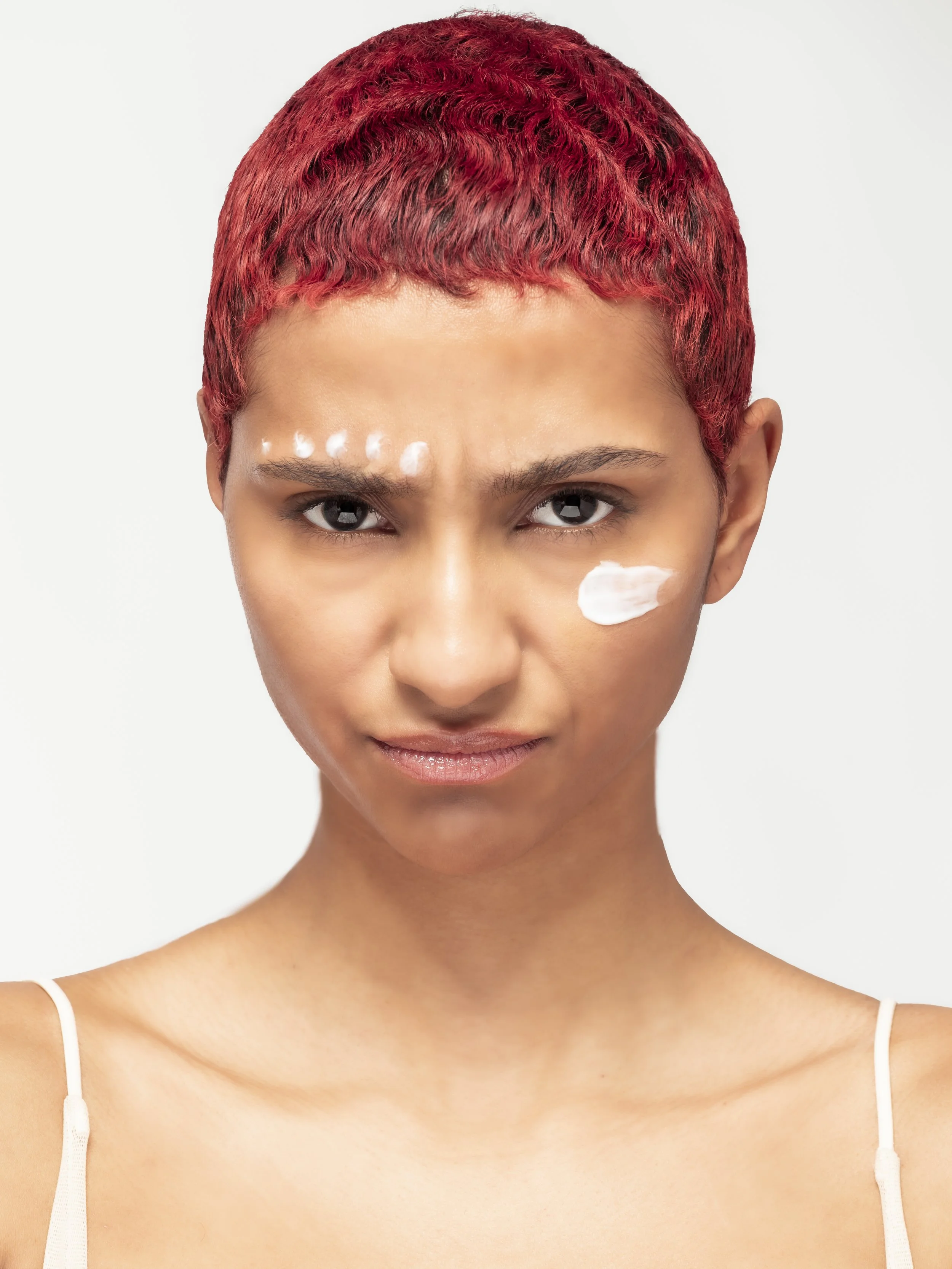 Jeune femme avec des cheveux courts rouges, regard déterminé, portant de la crème sur le visage, portant une robe blanche à fines bretelles, arrière blanc.