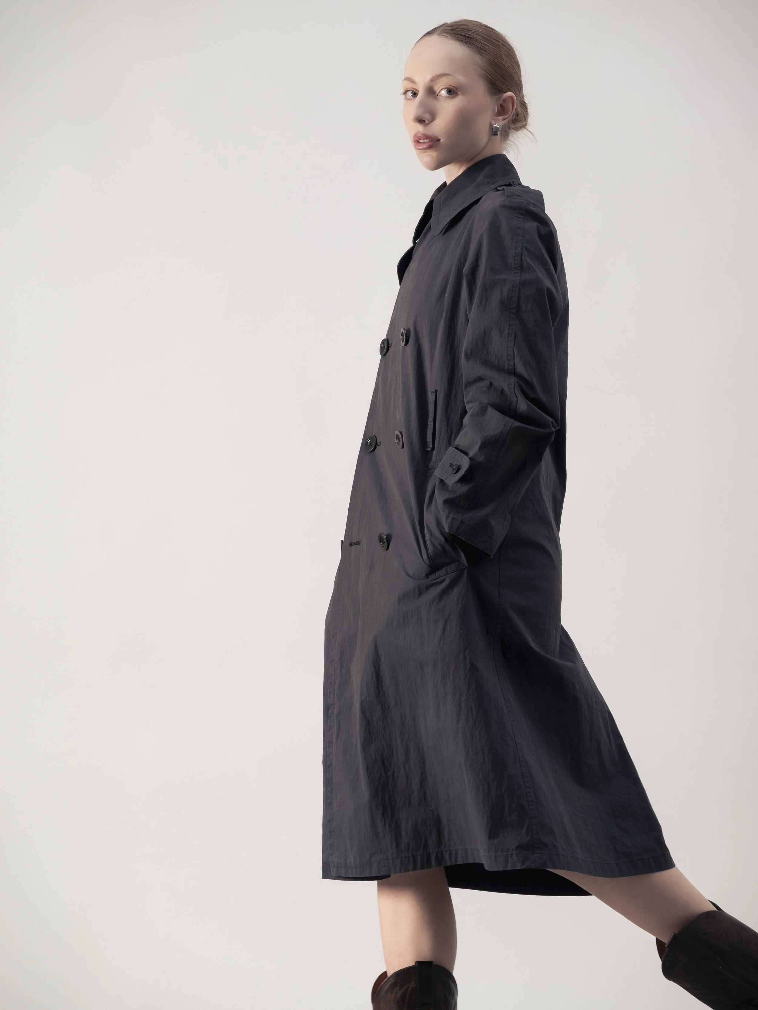 Femme portant un trench noir, avec une pose décontractée, contre un fond blanc.