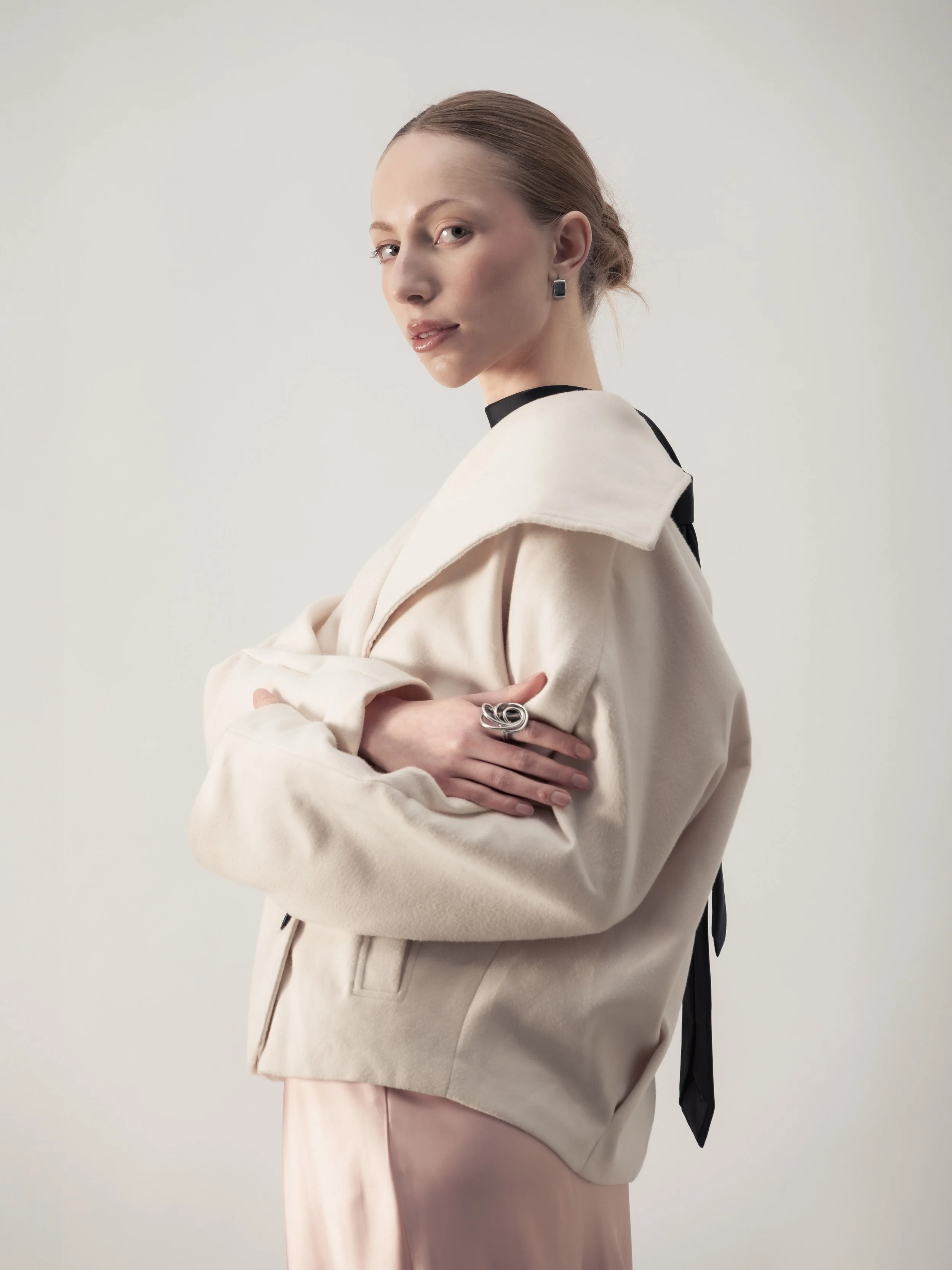 Une femme avec des cheveux roux tirés en arrière, portant une veste beige et une robe rose pâle, pose avec ses bras croisés contre un mur blanc, portant des boucles d'oreilles et une bague en argent.