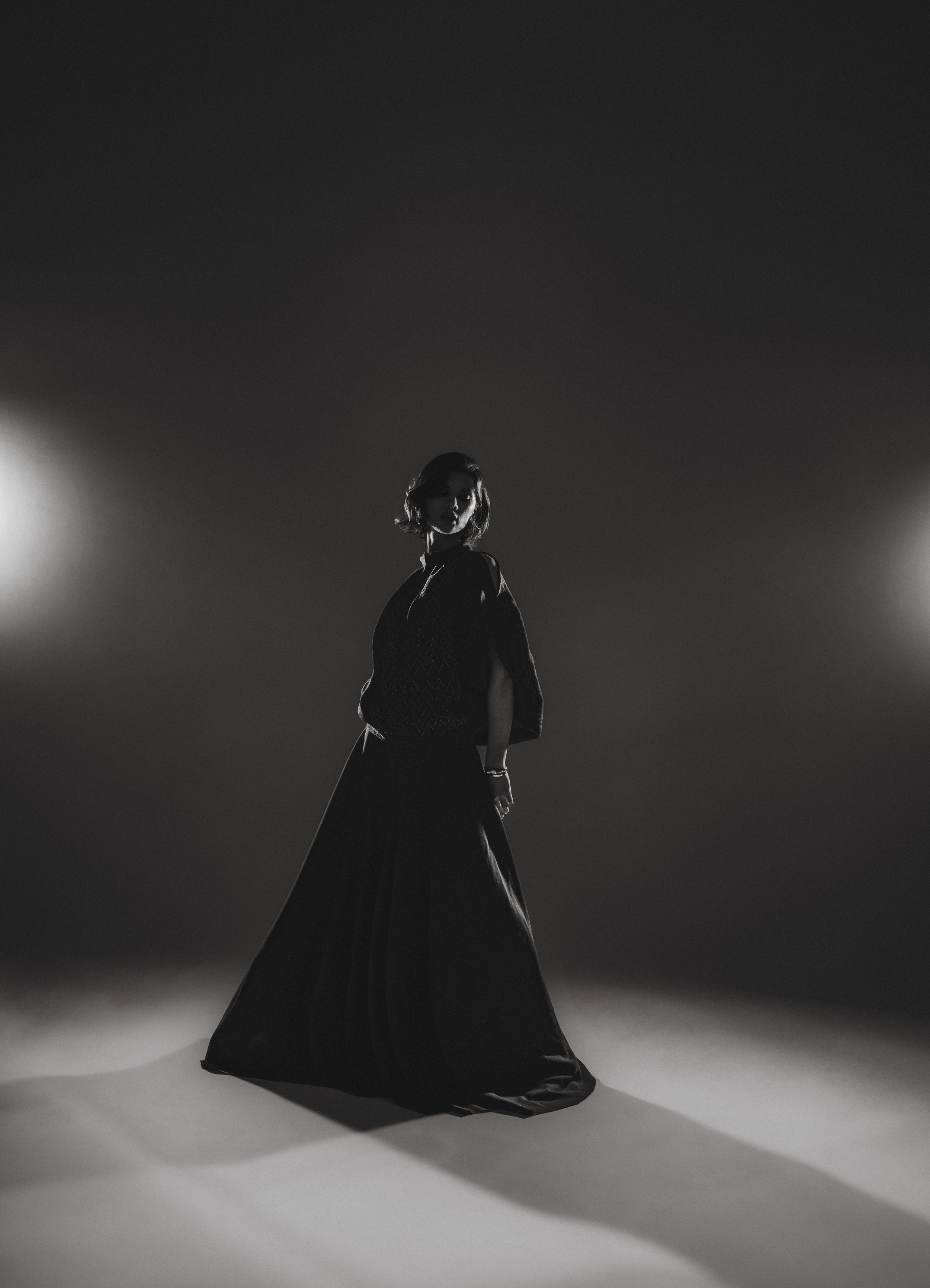 Une femme portant une robe longue noire, debout dans un environnement sombre avec deux sources de lumière derrière elle, créant un effet d'ombre et de lumière en silhouette.
