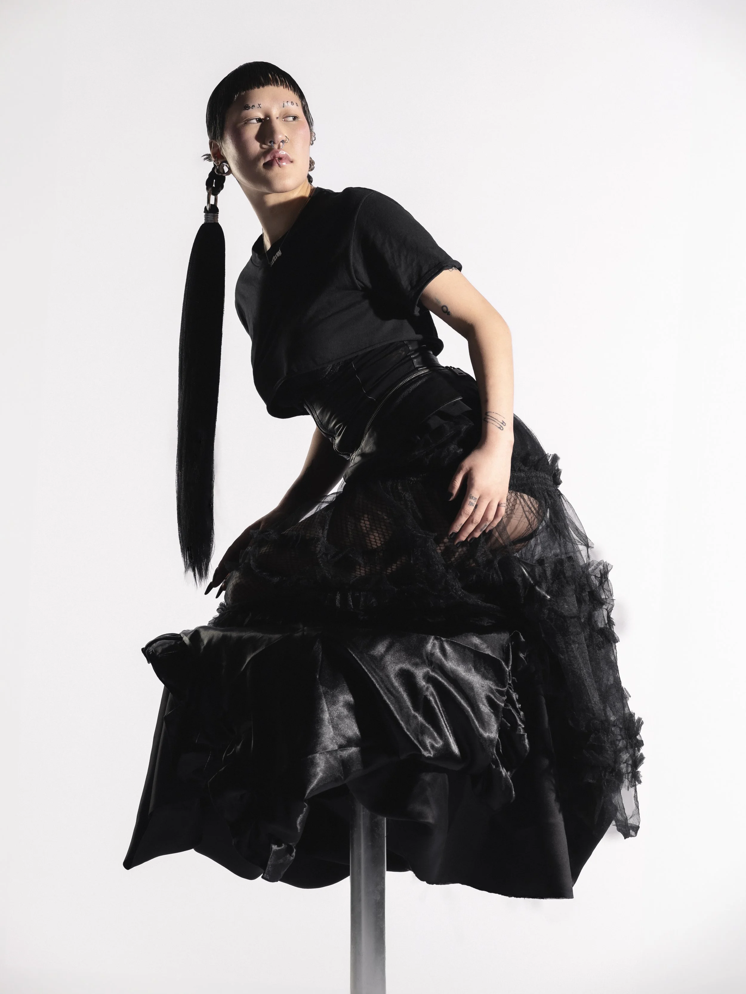 Une femme jeune avec un style gothique, portant des vêtements noirs, des bijoux, et des piercings, posant contre un fond blanc.